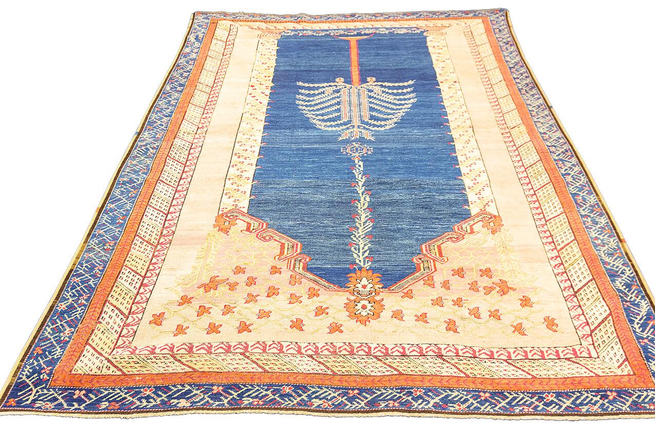 Immerse yourself in the enchanting world of our Antique Ghiordes Rug, a timeless masterpiece that boasts a serene blue field and a delicate pastel orange border. What makes this rug truly exceptional is the special tree motif carefully woven into