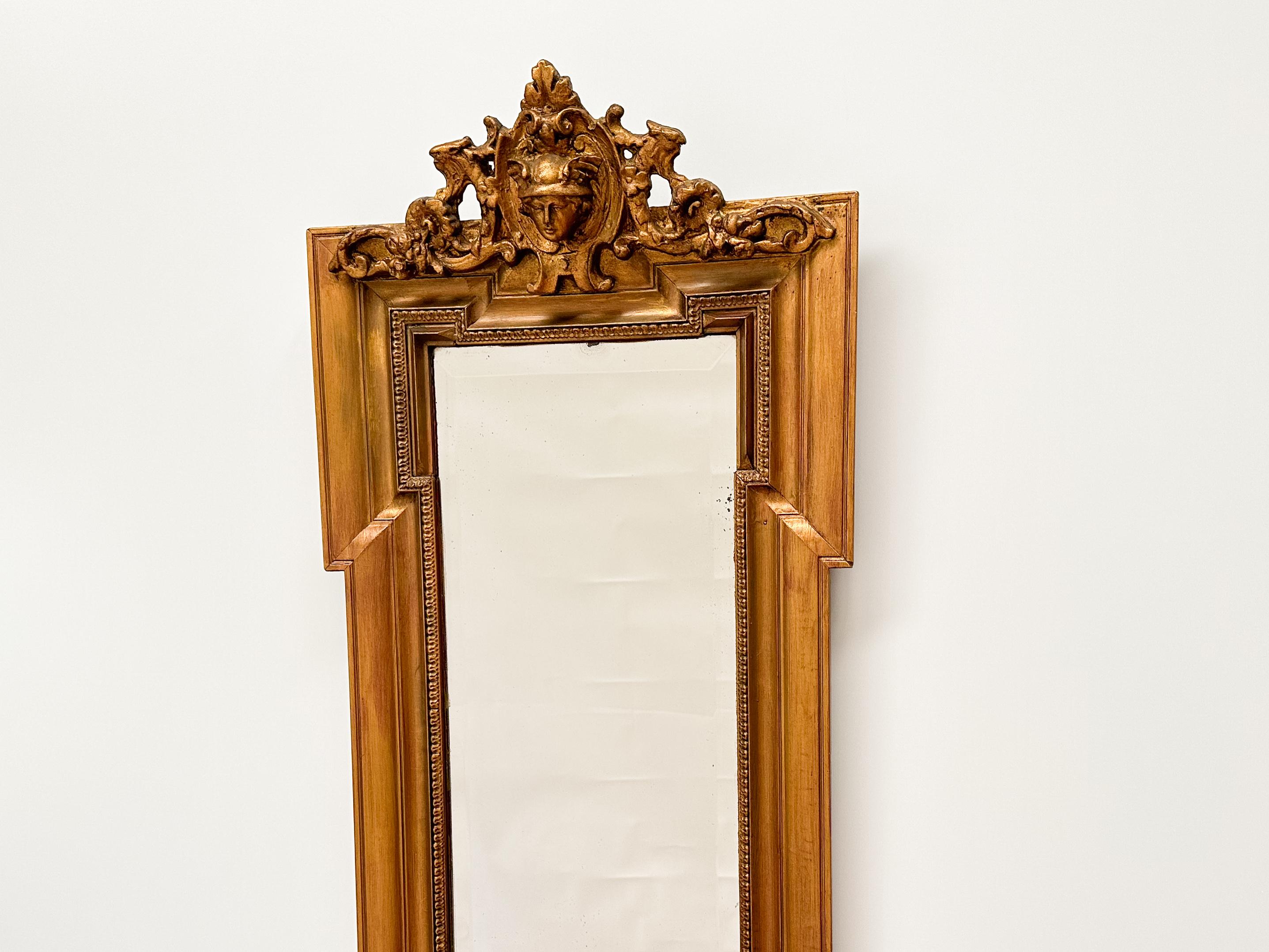 Elegant miroir mural ancien doté d'un cadre richement doré avec des détails complexes et un motif d'écusson classique. Le miroir est doté d'un verre biseauté d'origine, ce qui ajoute de la profondeur et du raffinement à son design intemporel. Le