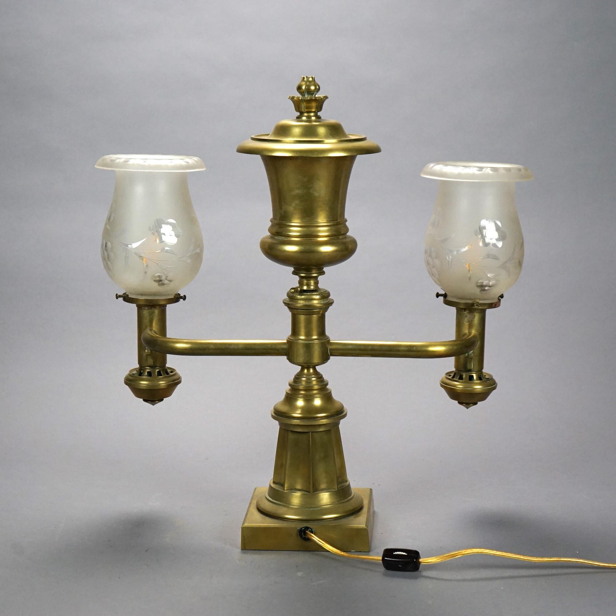 An antique argand table lamp offers cast brass and bronze base with font having floral finial and surmounting base with flared and faceted plinth with two arms terminating in lights having frosted glass shades with floral decoration, electrified,