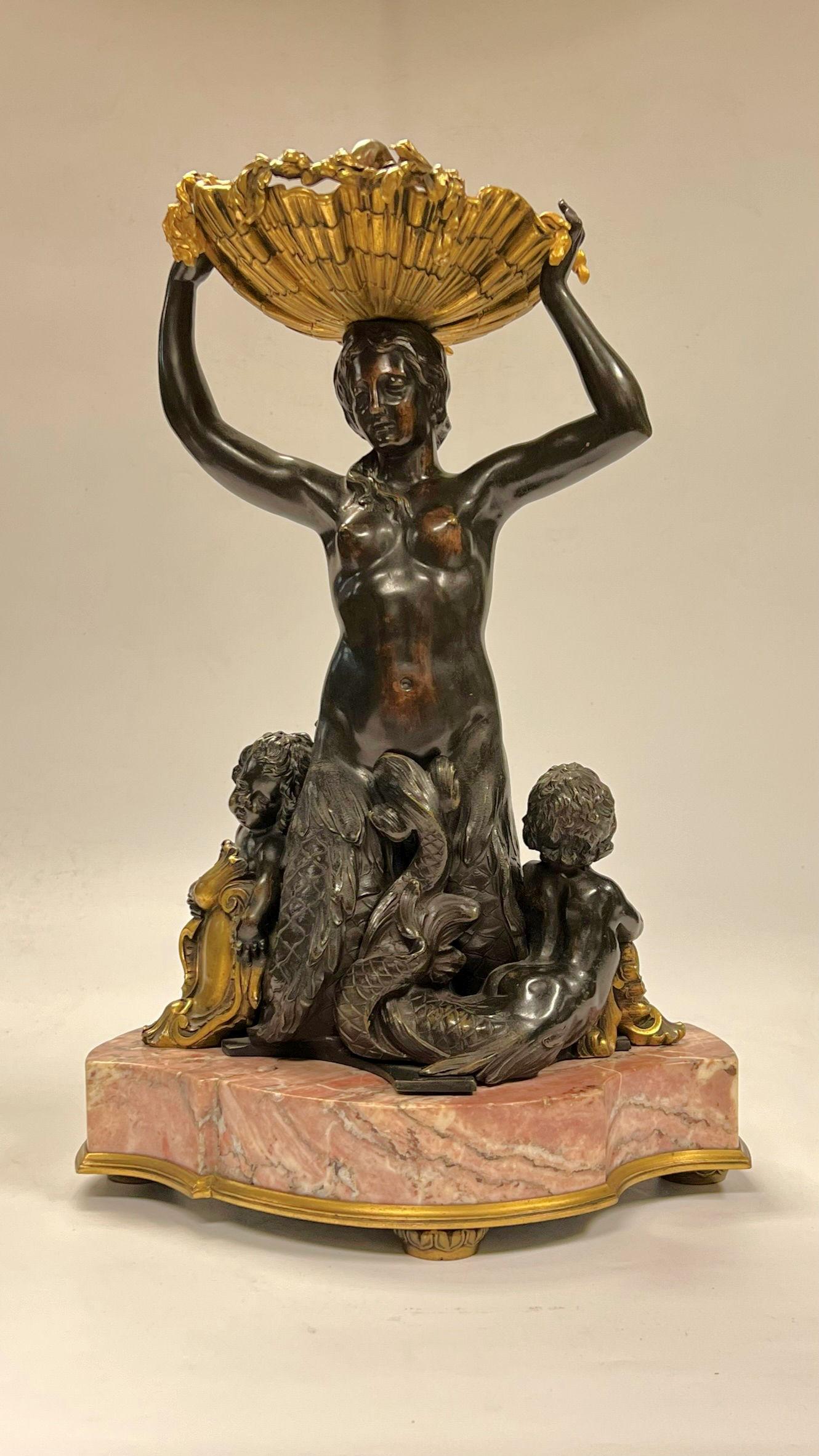 Gilt and patinated bronze centerpiece depicting a lovely mermaid supporting a seashell with stylized dolphin with cherubs (putti) at the base, mounted on a variegated pink marble pedestal and raised on gilt bronze feet.

This model appears at least