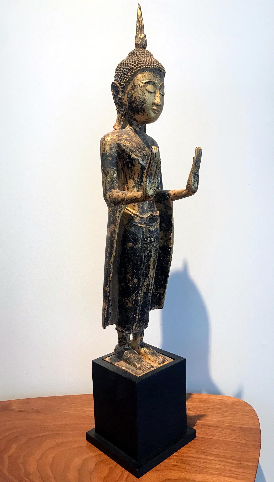Ein zart geschnitzter Holzbuddha in aufrechter Haltung mit doppelter Abhaya Mudra. Die Statue weist eine vergoldete Oberfläche mit schöner Patina und leichten Gebrauchsspuren auf. Das Gesicht wurde mit einem heiteren Ausdruck mit niedergeschlagenen