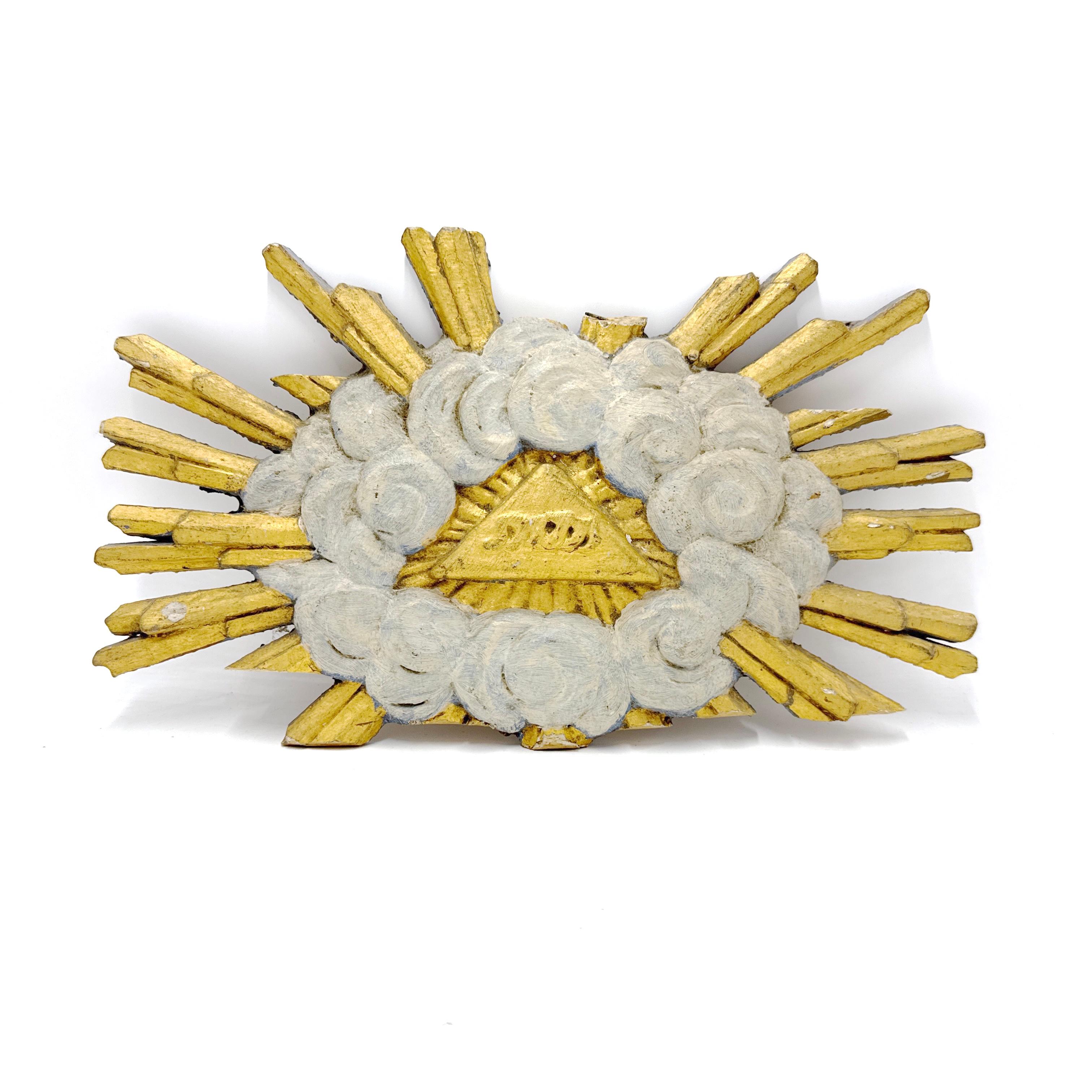 Un'autentica scultura ecclesiastica in legno dorato con il Tetragramma (יהוה) all'interno di un triangolo luminoso, che simboleggia il nome divino di Dio. Circondato da nuvole vorticose intagliate a mano e raggi di sole, questo pezzo rappresenta la