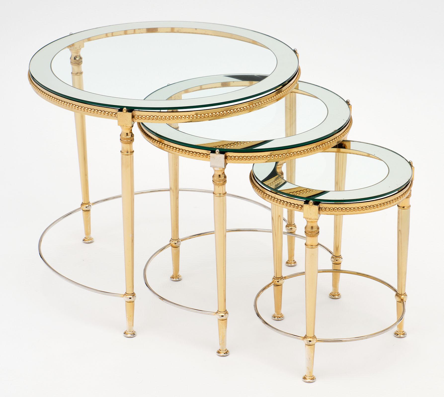 Classic set of glass and brass antique nesting tables made of gilded brass. Each features the original round glass tops with mirrored edges. The measurements listed are for the largest table. Please find dimensions for the smaller two