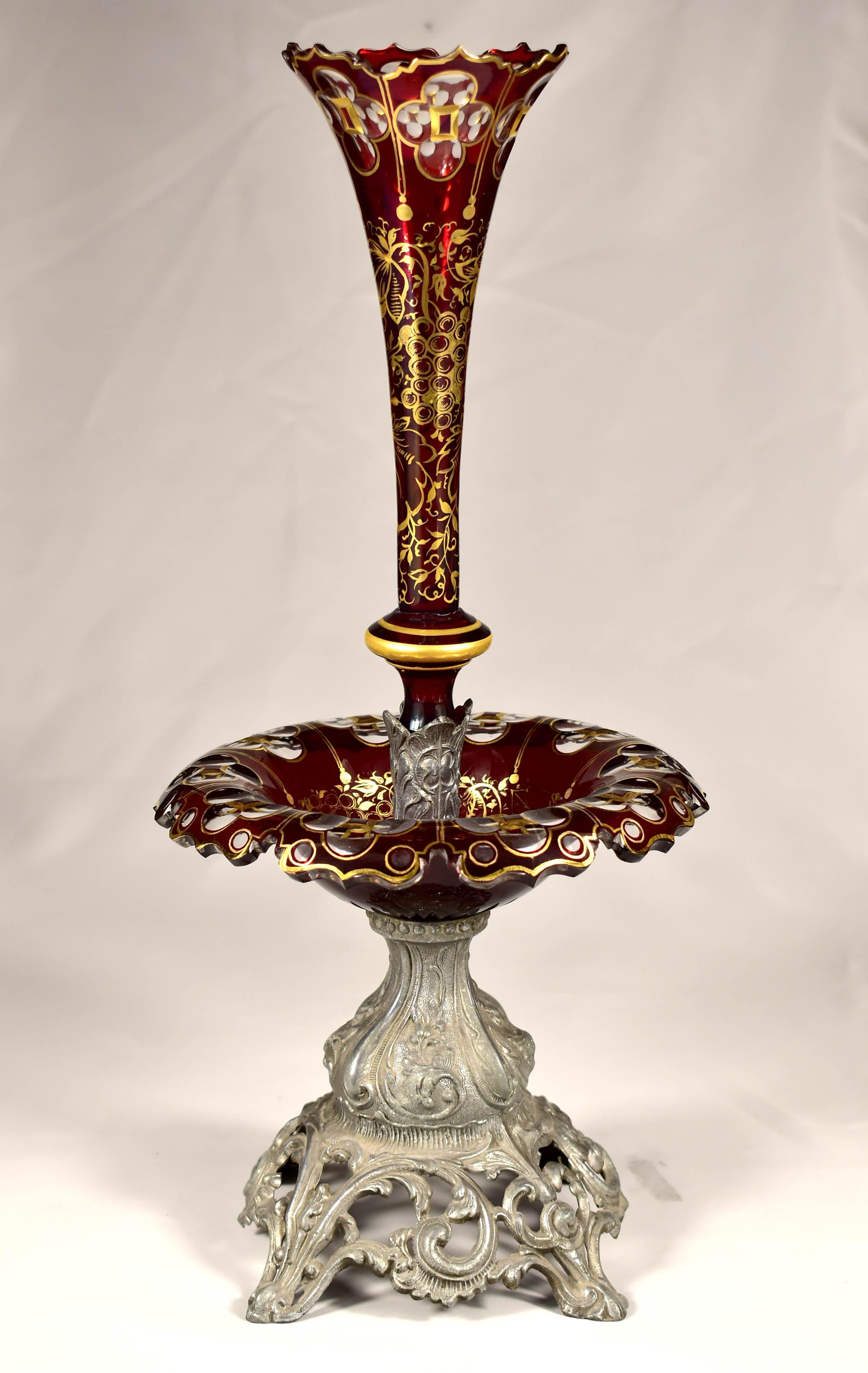 The body of the bowl was made of tin alloy. A wide glass bowl or plate was used to display fruit or other goodies. The top glass in the shape of a tall, narrow flower serves as a vase. Both glass pieces have cut edges. The glass parts are made of