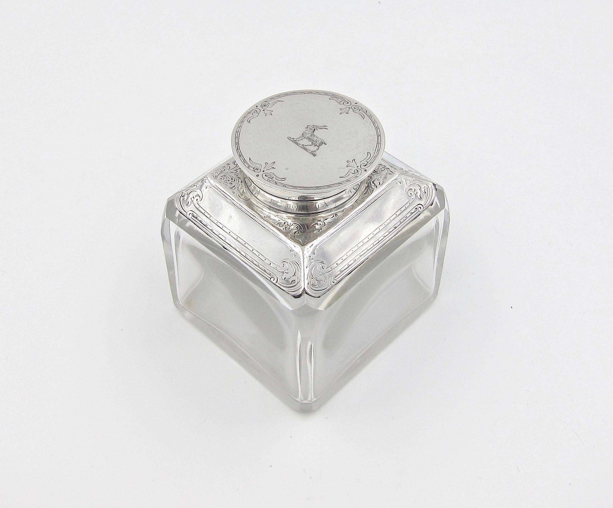 An antique glass inkwell topped with a sterling silver hinged lid and collar, made in England in 1894. This Victorian desk accessory was crafted from heavy glass with canted corners. The hinged lid and silver mounts were hand-engraved with delicate