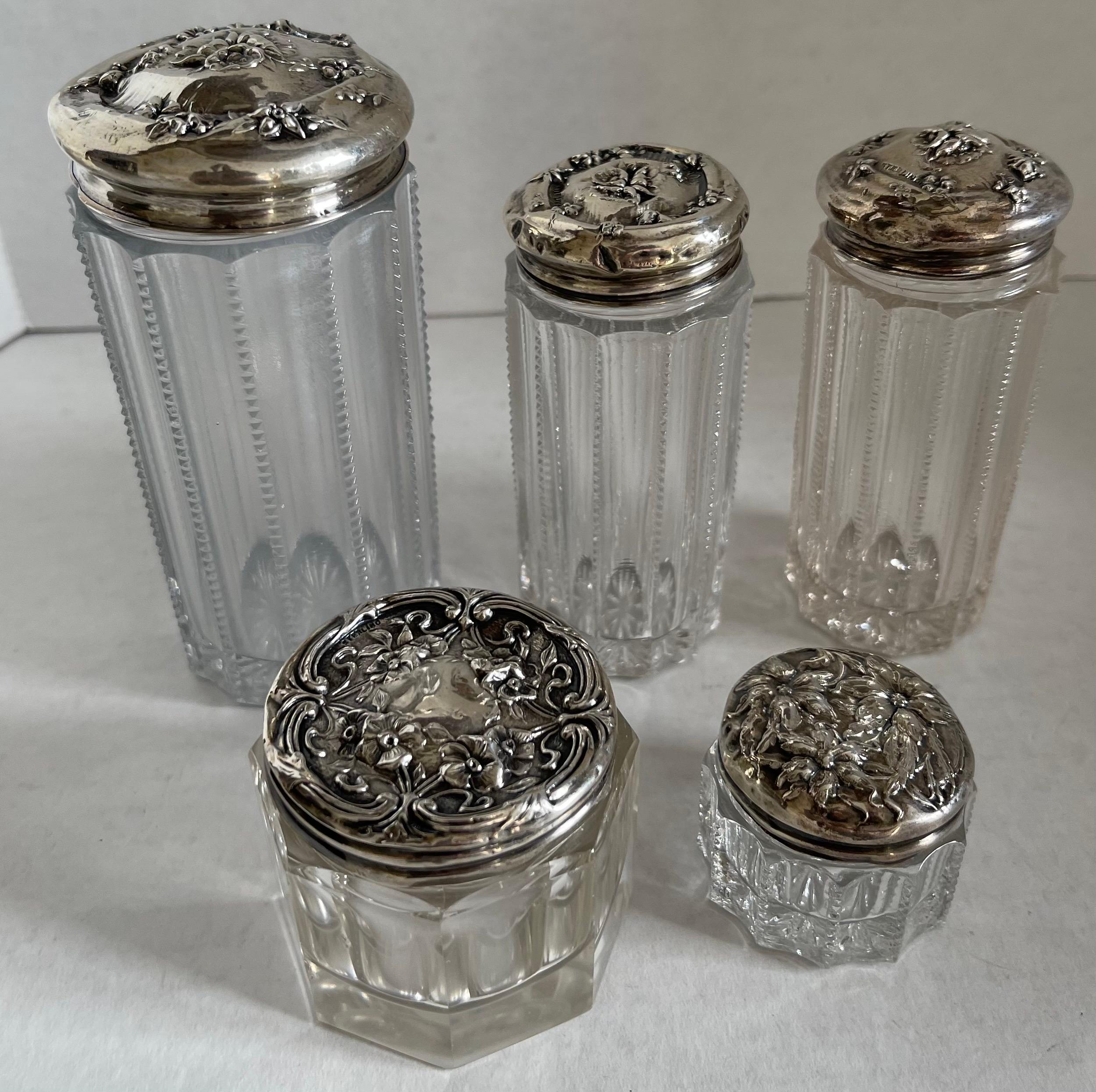 Set of five antique sterling silver and glass vanity jars. Various repoussè floral designs. each sterling lid is stamped sterling. 
Large jar measures: 4.5” tall x 2” wide. 2 medium jars each measure: 3.5” tall x 2” wide. Small jar measures: 2” tall