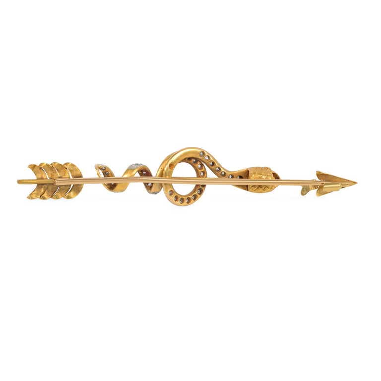 An antique emerald, diamond, and gold brooch in the form of an arrow with a coiled serpent with a pavé diamond body, and the head with ruby eyes and centered by an emerald, in 18k and platinum. French import.