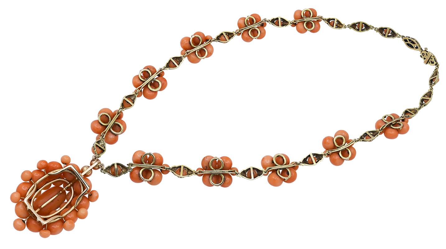 The oval pendant set with polished pale orange coral buttons suspended from a decorative yellow gold fancy link chain set at intervals with ten smaller clusters of similar pale orange coral buttons, measures app. 20 inches long, the drop measures