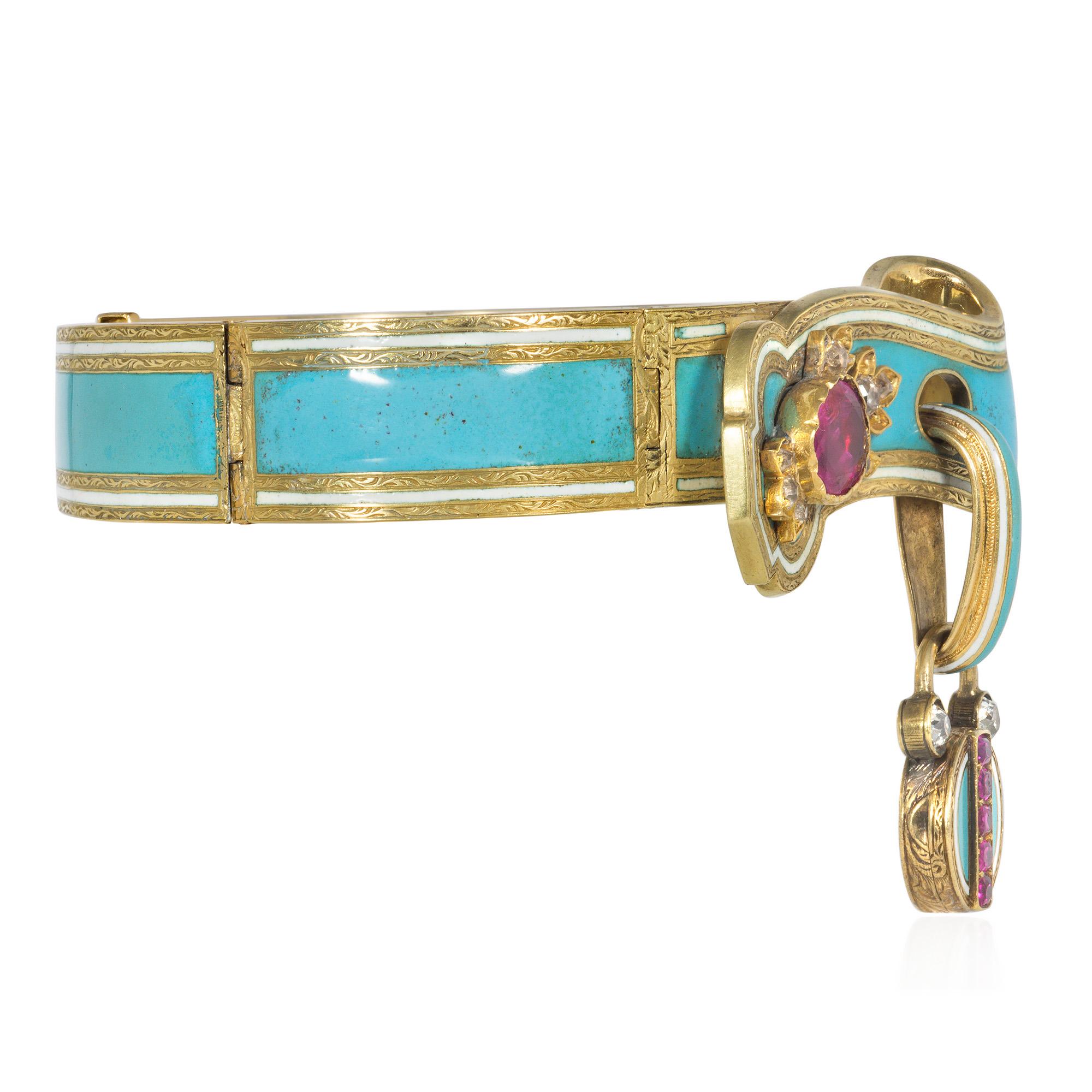 An antique early Victorian gold and enamel hinged bangle bracelet designed as a scrolling ribbon garter in chased gold and turquoise and white enamel, with a large ruby flanked by foliate motifs accented with diamonds, suspending a similarly