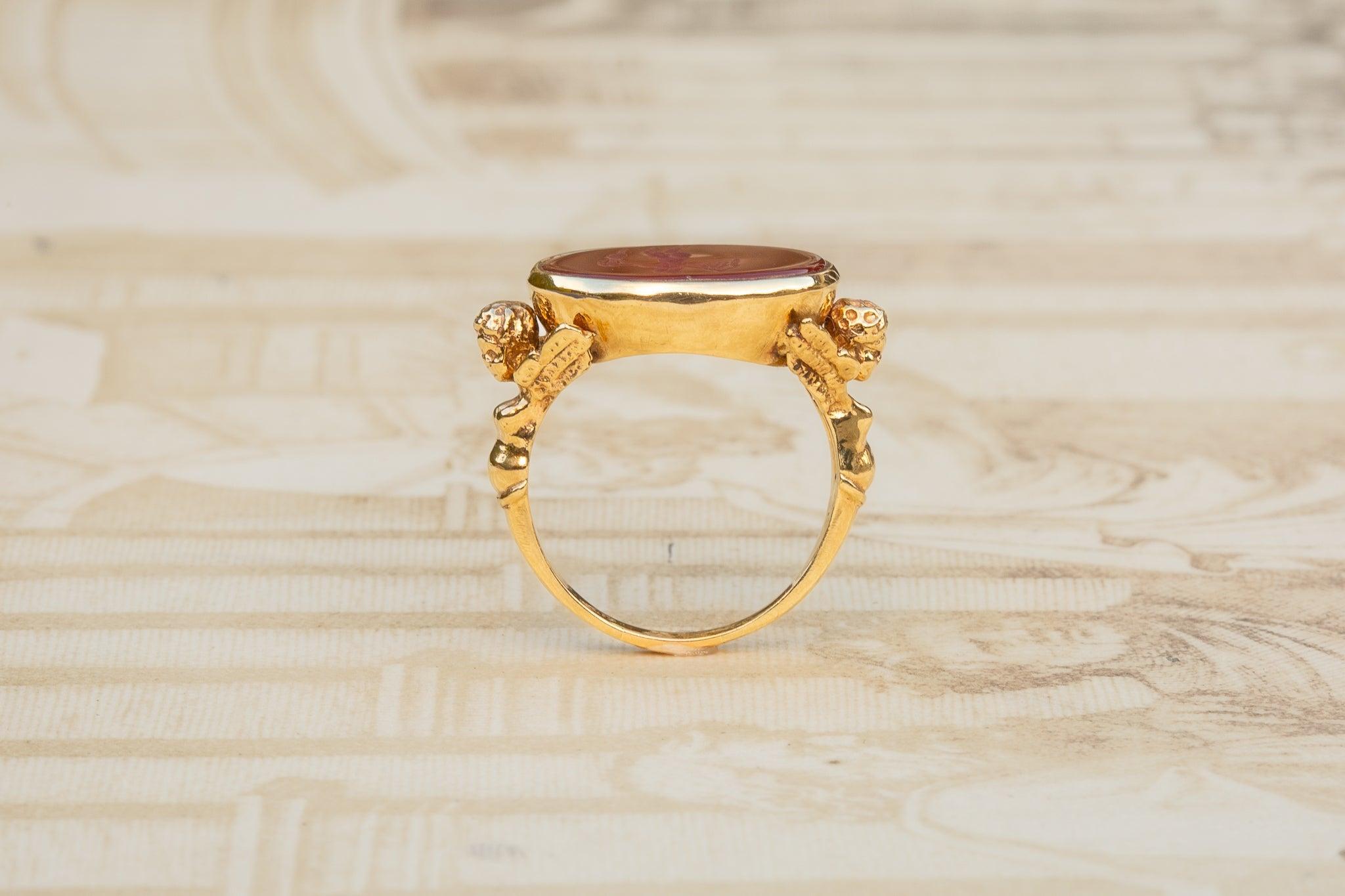 A striking gold ring, dating from the early 20th century. The figural shoulders are adorned with angelic putti motifs, delicately sculpted to depict Eros, the Greek God of love and desire, who is often associated with Cupid in Roman mythology. These