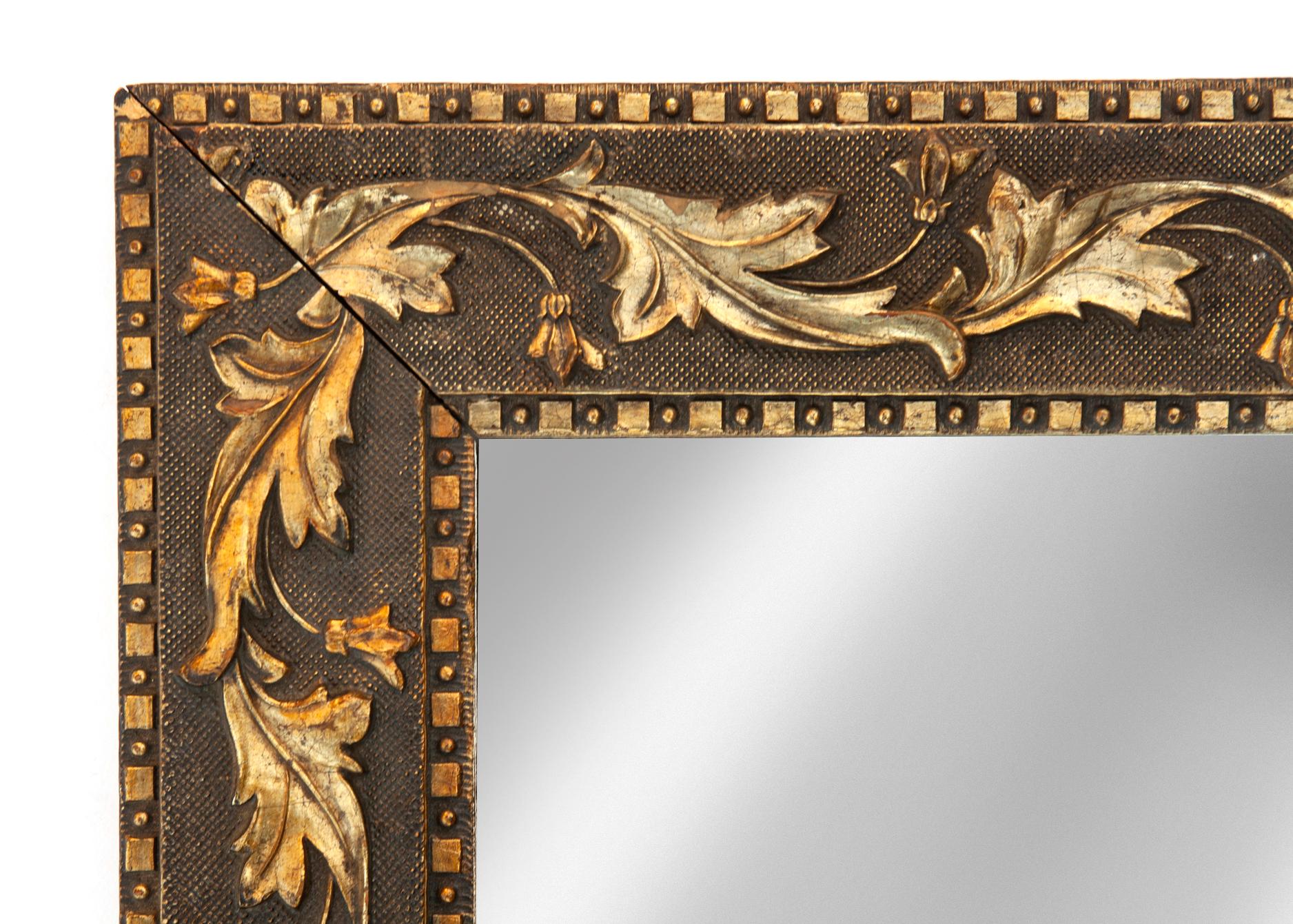 This small mirror is embellished with burnished gold swirling leaves on a dark bronze textured background. The exterior and interior border is comprised of a geometric pattern of small squares and dots. 
This mirror is a rich & interesting accent