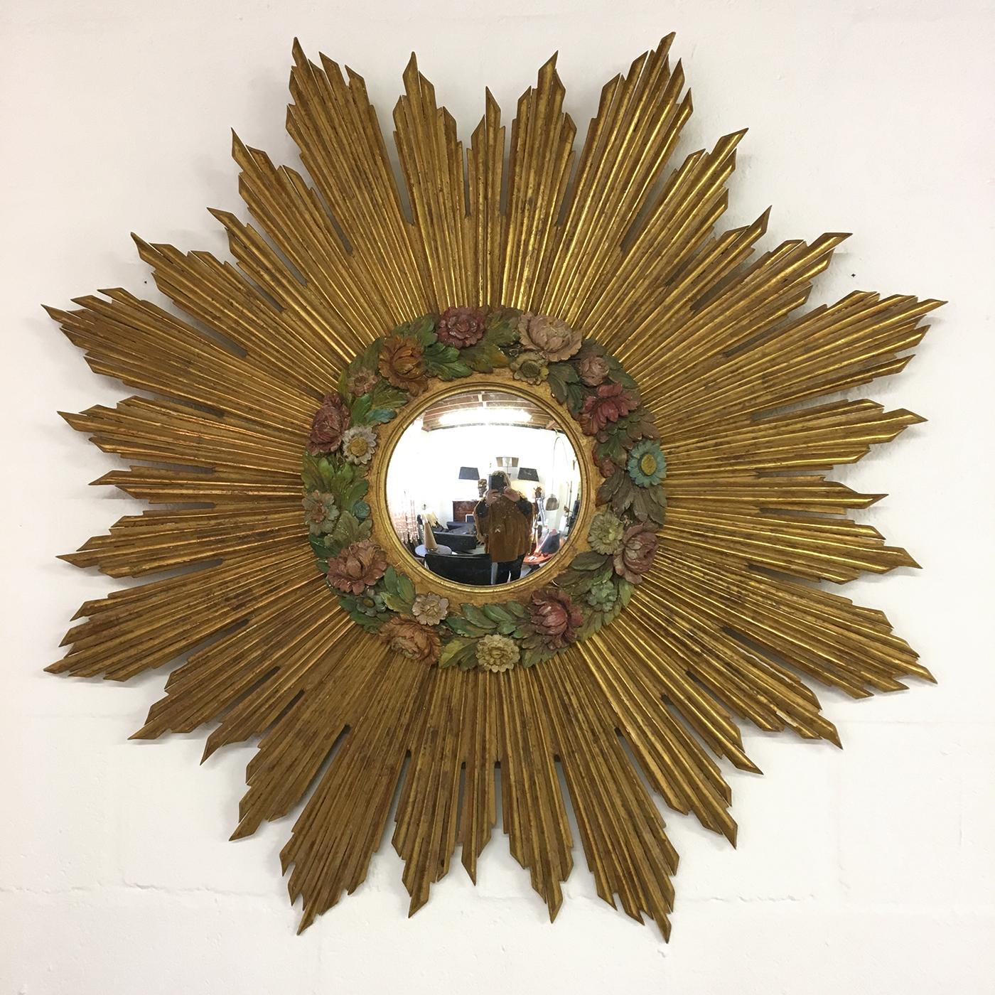 A magnificent and very large early to mid-20th century French gilded and carved wood sunburst with a convex mirror, featuring a beautiful carved and highly decorative wood polychrome Barbola wreath of flowers – which was a trend in the