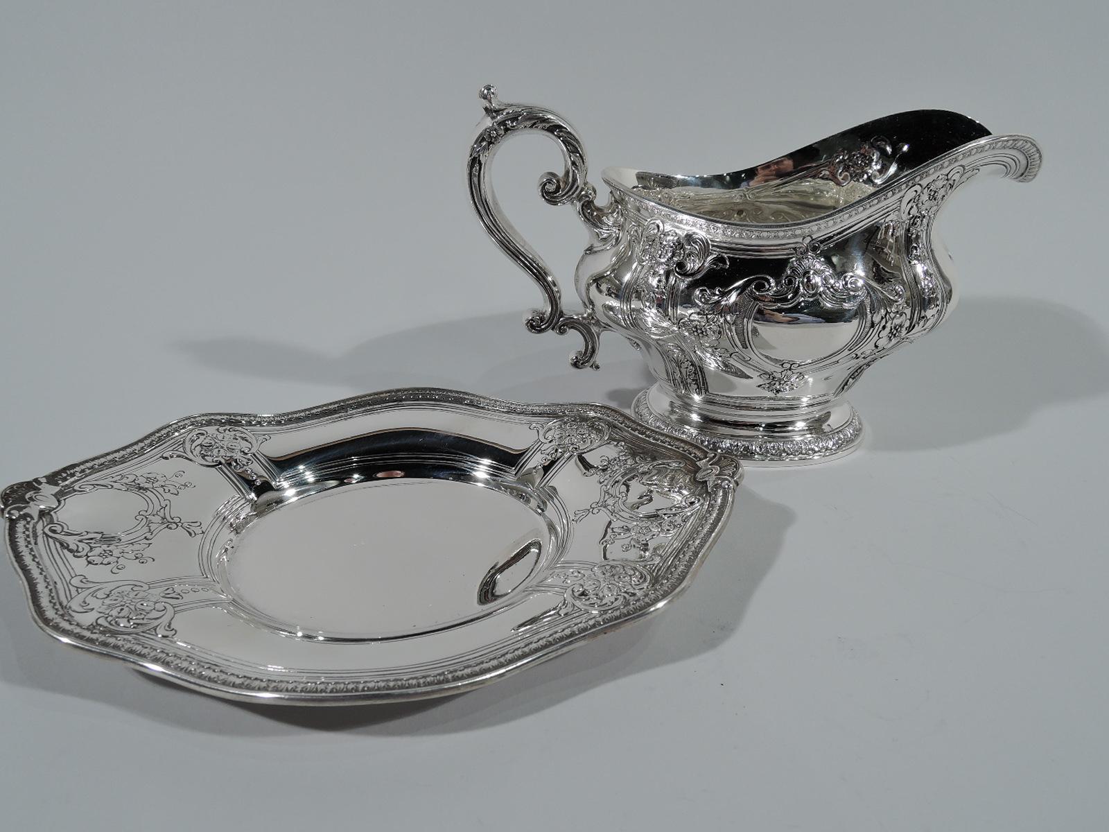 Fancy sterling silver gravy boat on stand in Gregorian pattern. Made by Gorham in Providence. Boat: Bellied with helmet mouth, leaf-capped s-scroll handle, and raised oval foot. Stand: Shaped with oval well. Pendant flowers, shells, and armorial