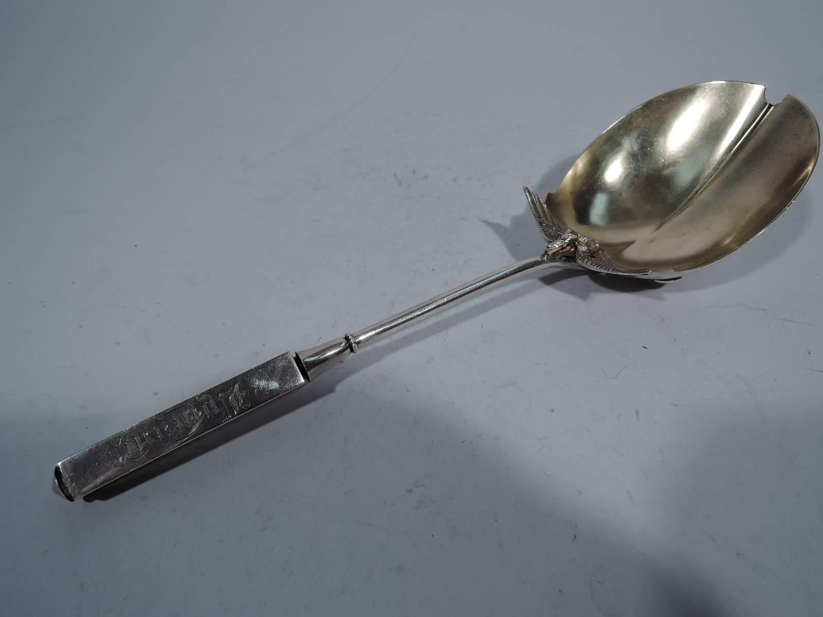 Sterling silver berry spoon in Isis pattern. Made by Gorham in Providence. Narrow cylindrical stem with split volute scrolls mounted to deep round bow. Applied to bowl are coiled serpent and wings. Bowl interior and ornament gilt. Rectilinear