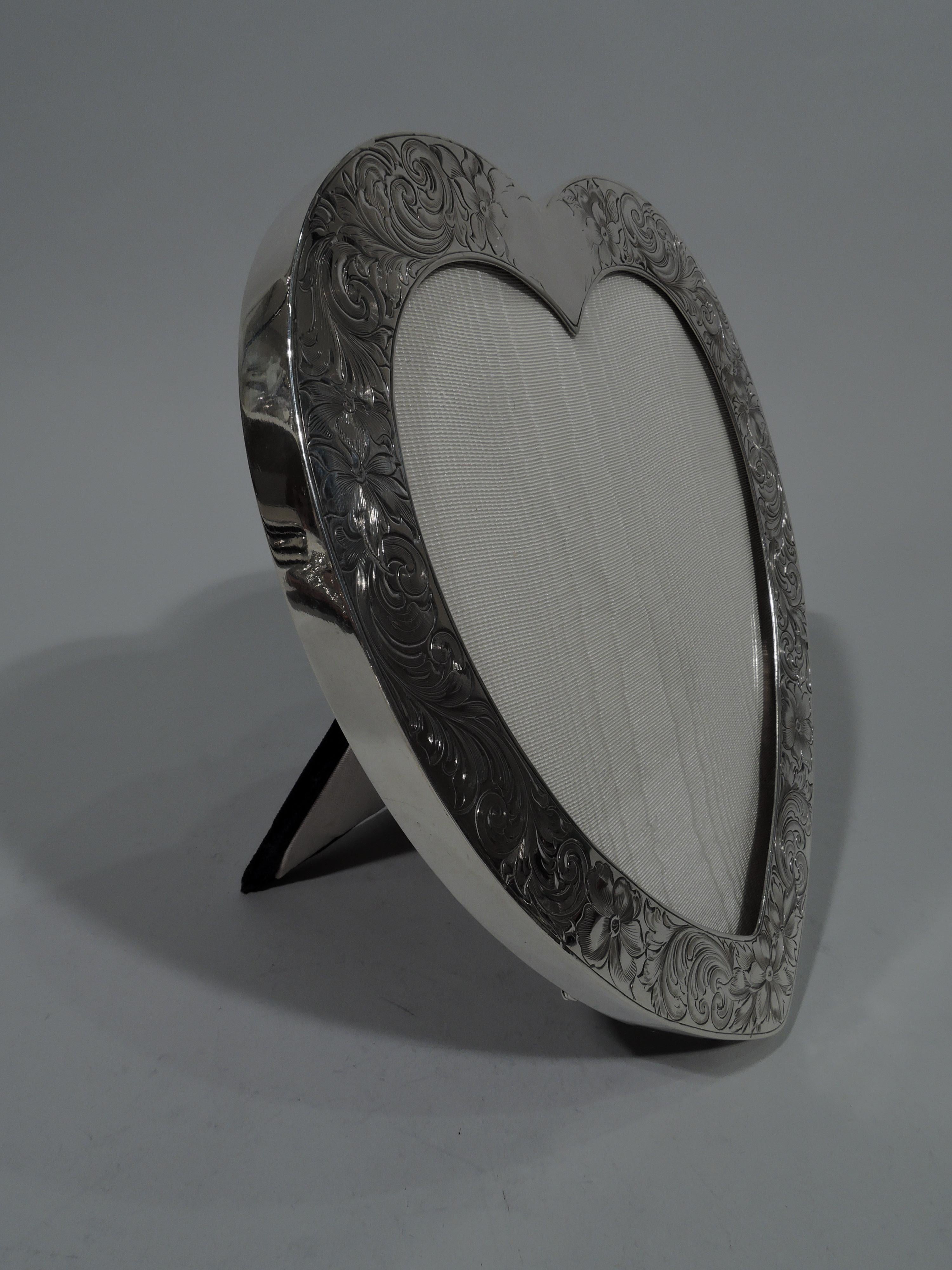 Romantic sterling silver heart frame. Made by Gorham in Providence, circa 1910. Flat surround with engraved scrolls and flowers. Shaped cartouche (vacant) at top. With glass, silk lining, and velvet back and hinged support. Fully marked including