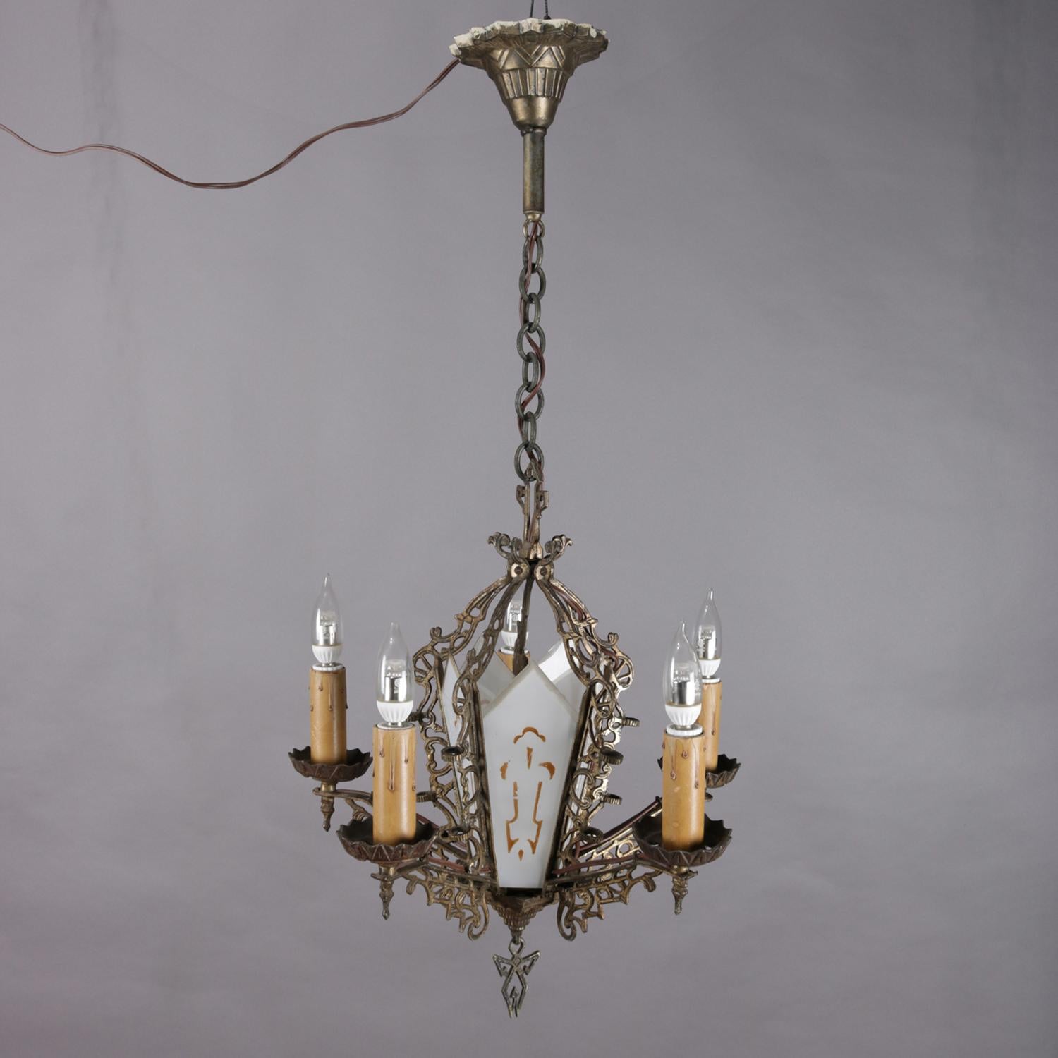 Antique Gothic style chandelier features pierced bronze frame housing cathedral arch form gilt stenciled frosted glass panels surrounding internal light and having five arms terminating in candle lights with gilt drips and central stylized angel