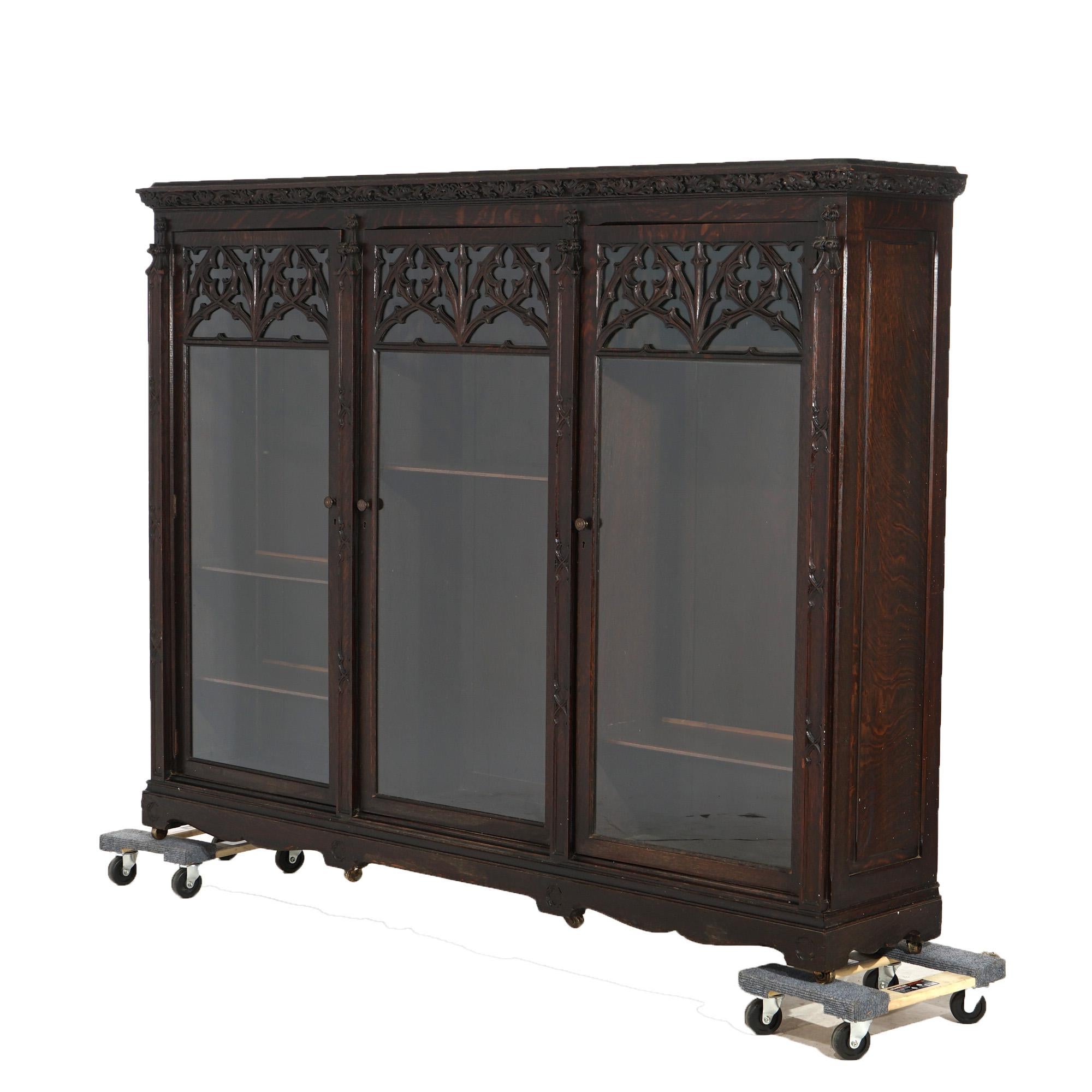 An antique Gothic Revival bookcase offers deeply striated quarter sawn oak construction with deeply carved acanthus frieze surmounting case having three glass doors with carved quatrefoil tracery elements and opening to divided shelved interior;