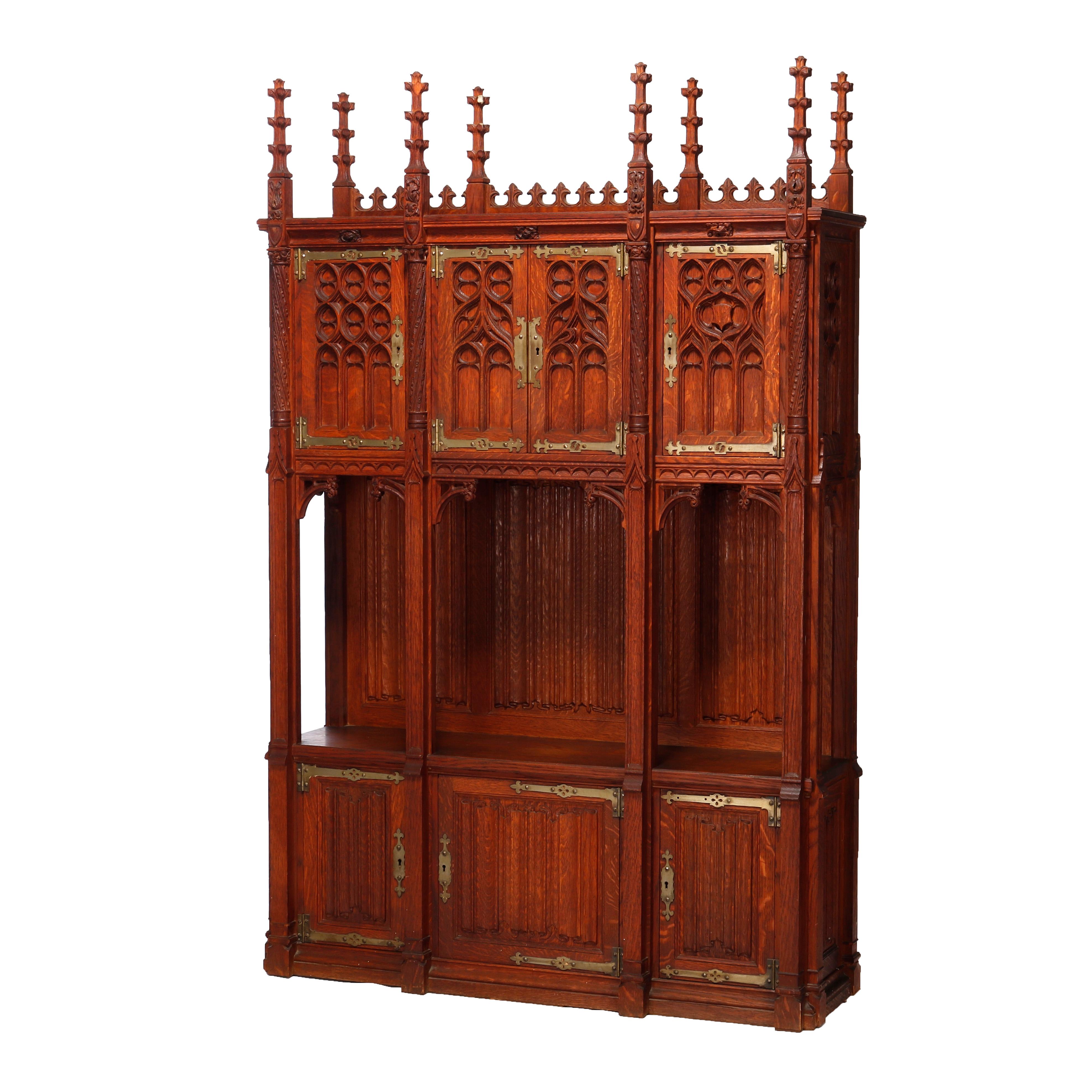 An antique Gothic Revival cabinet in the manner of Kimbel and Cabus offers quarter sawn oak construction with upper having spires with stylized foliate elements over upper with heavily carved doors having strap hinges and lower cabinet having