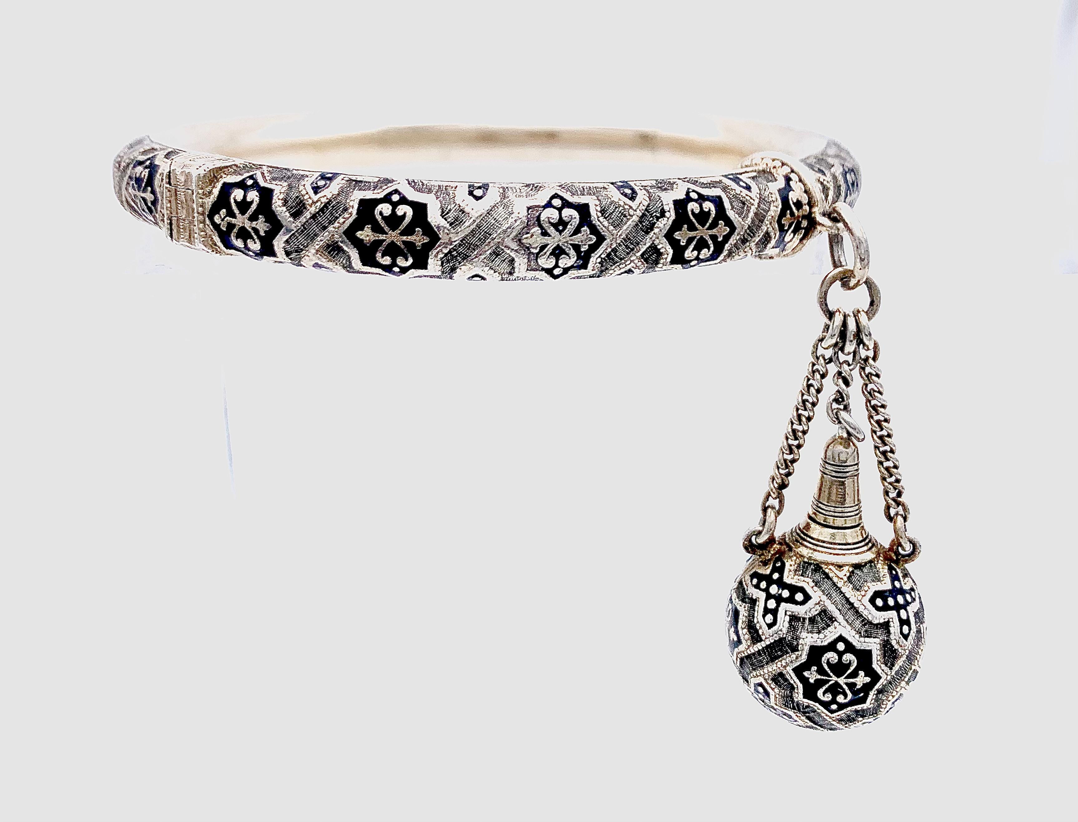 This highly unusual silver bangle has a miniature perfume bottle suspended from it. Bracelet and bottle are finely engraved with geometric ornaments in the gothic revival taste. The ornaments are highlighted with black enamel.