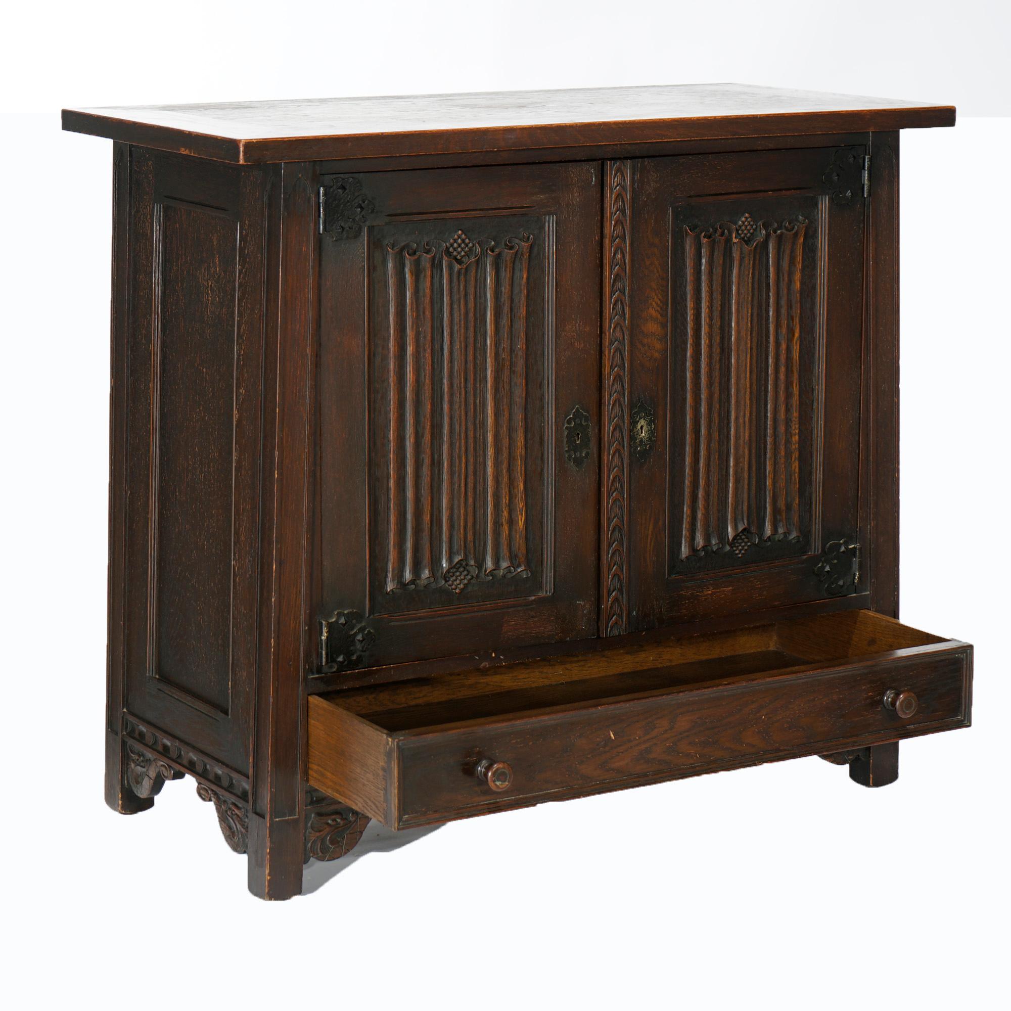 An antique Gothic style credenza in the manner of Kittinger offers English oak construction with flared case having double doors with carved reserves opening to shelved interior with slide-out trays over single long drawer and raised on feet bracket