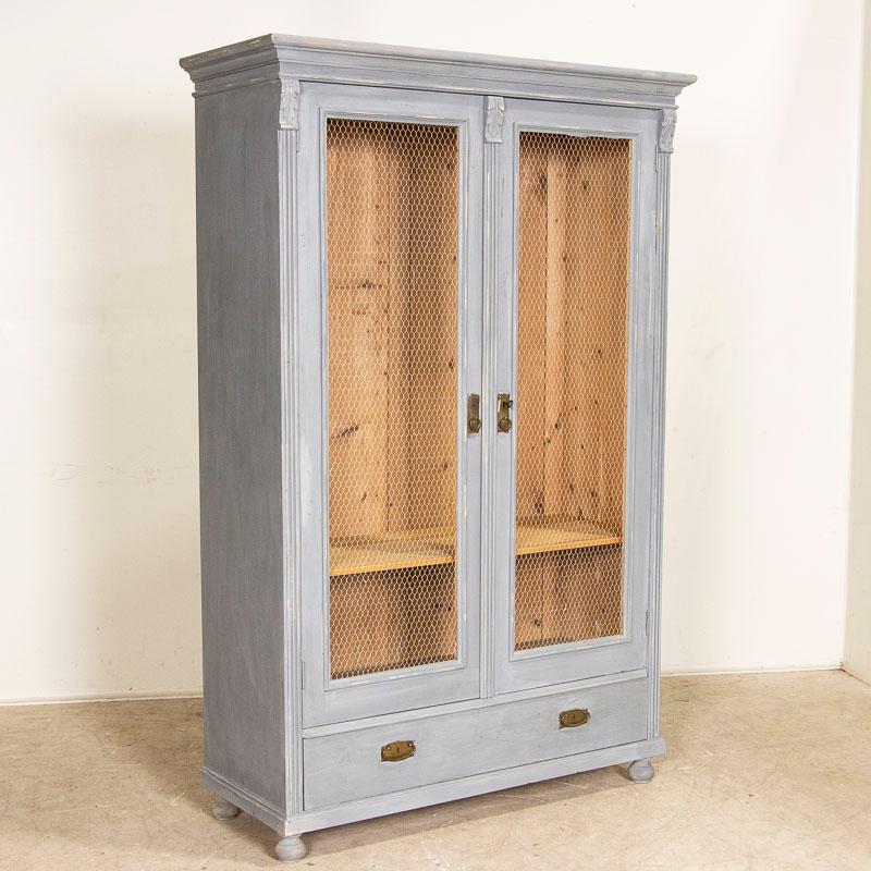 This antique gray painted 2 door cabinet with lower drawer has had the glass replaced with wire mesh giving the bookcase a more open and accessible feel. Where the (newer) grayish blue paint has been scraped there are hints of natural pine showing