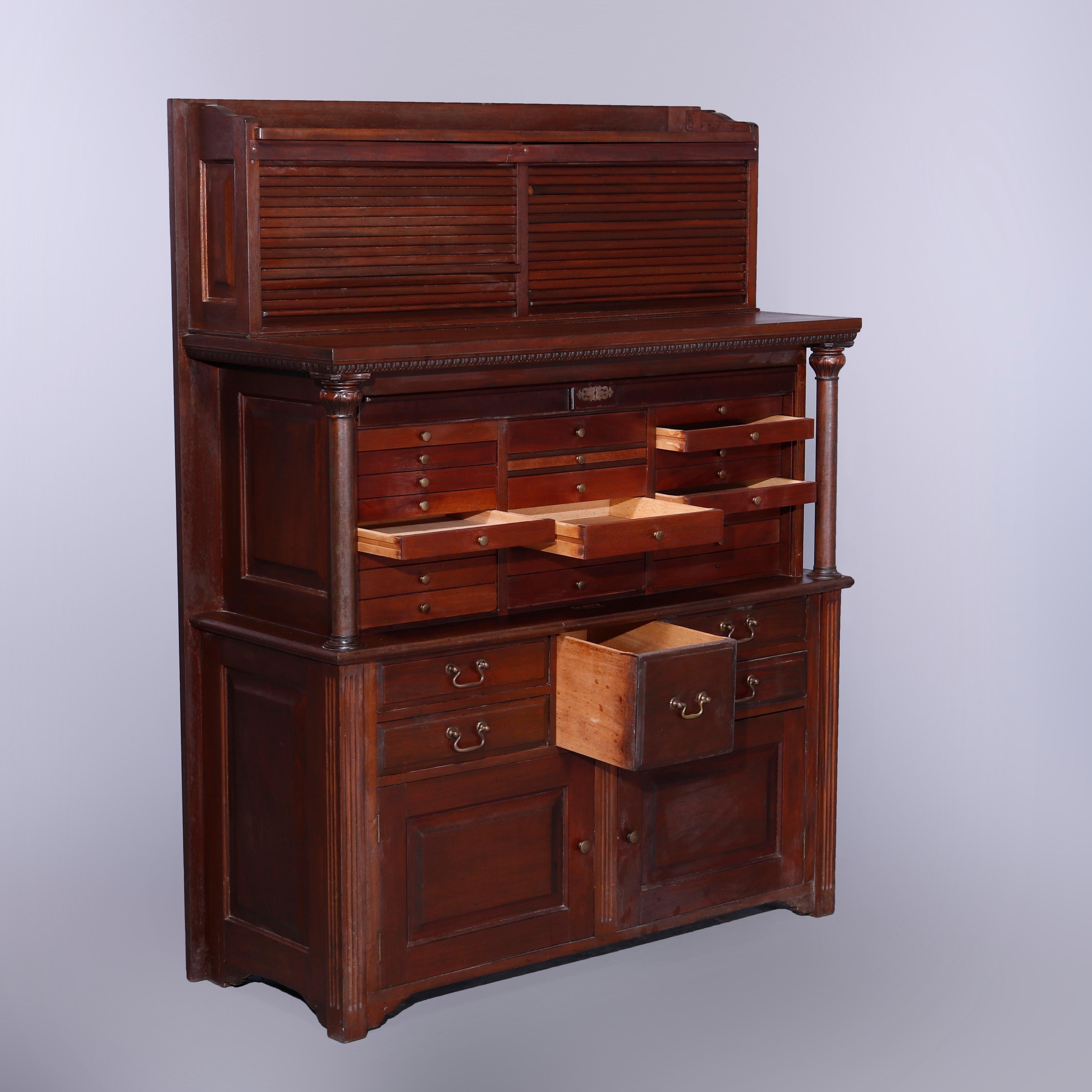 An antique American Empire dental cabinet offers raised panel mahogany construction with upper having tambour doors raising to reveal compartmentalized storage over center section with twenty-two small drawers and flanking Grecian Doric Column