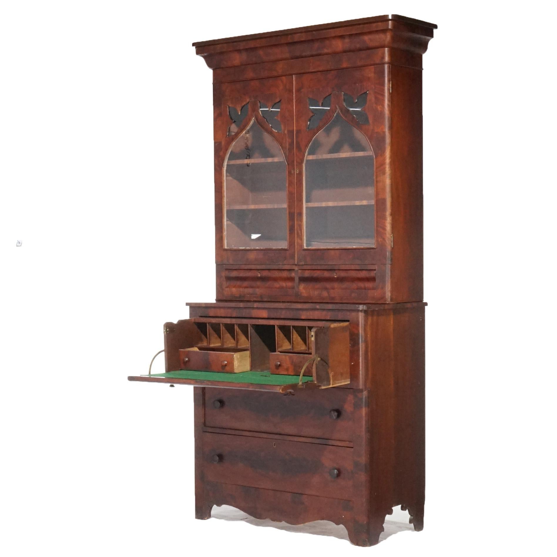 An antique American Empire secretary in the manner of Quervelle offers deeply striated flame mahogany construction with Greco-Roman features having upper bookcase with double glass doors with arch form overlay surmounting lower case with drop front