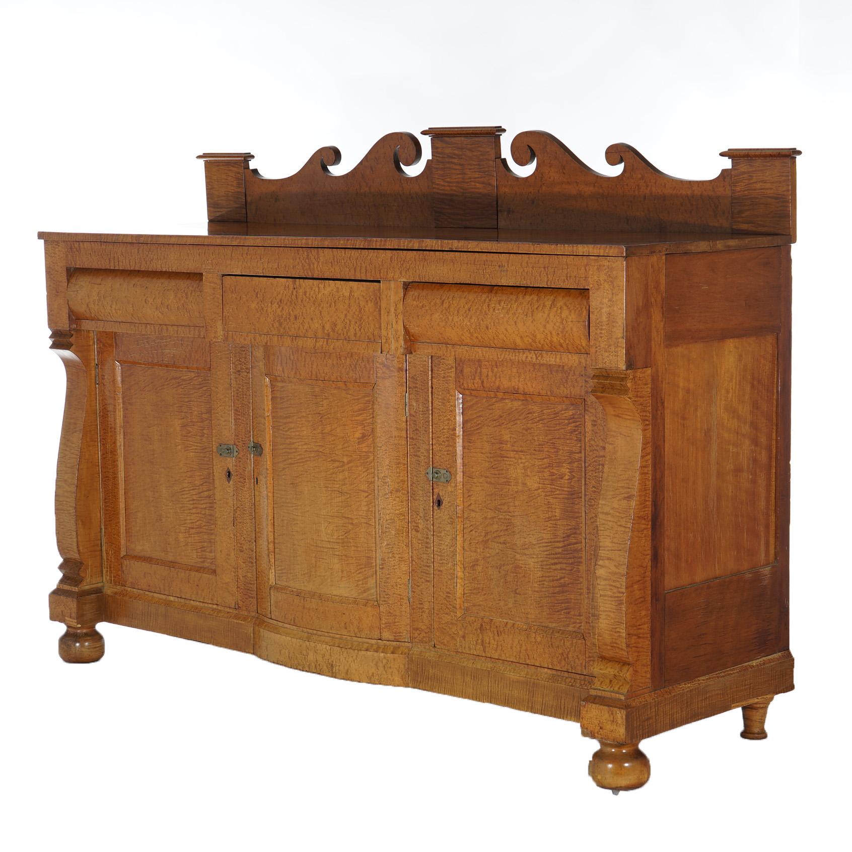An antique Greco American Empire sideboard offers tiger maple construction with scroll wave form backsplash over case with three upper drawers over three blind cabinets opening to shelved interiors, flanked by scroll form supports, raised on bun