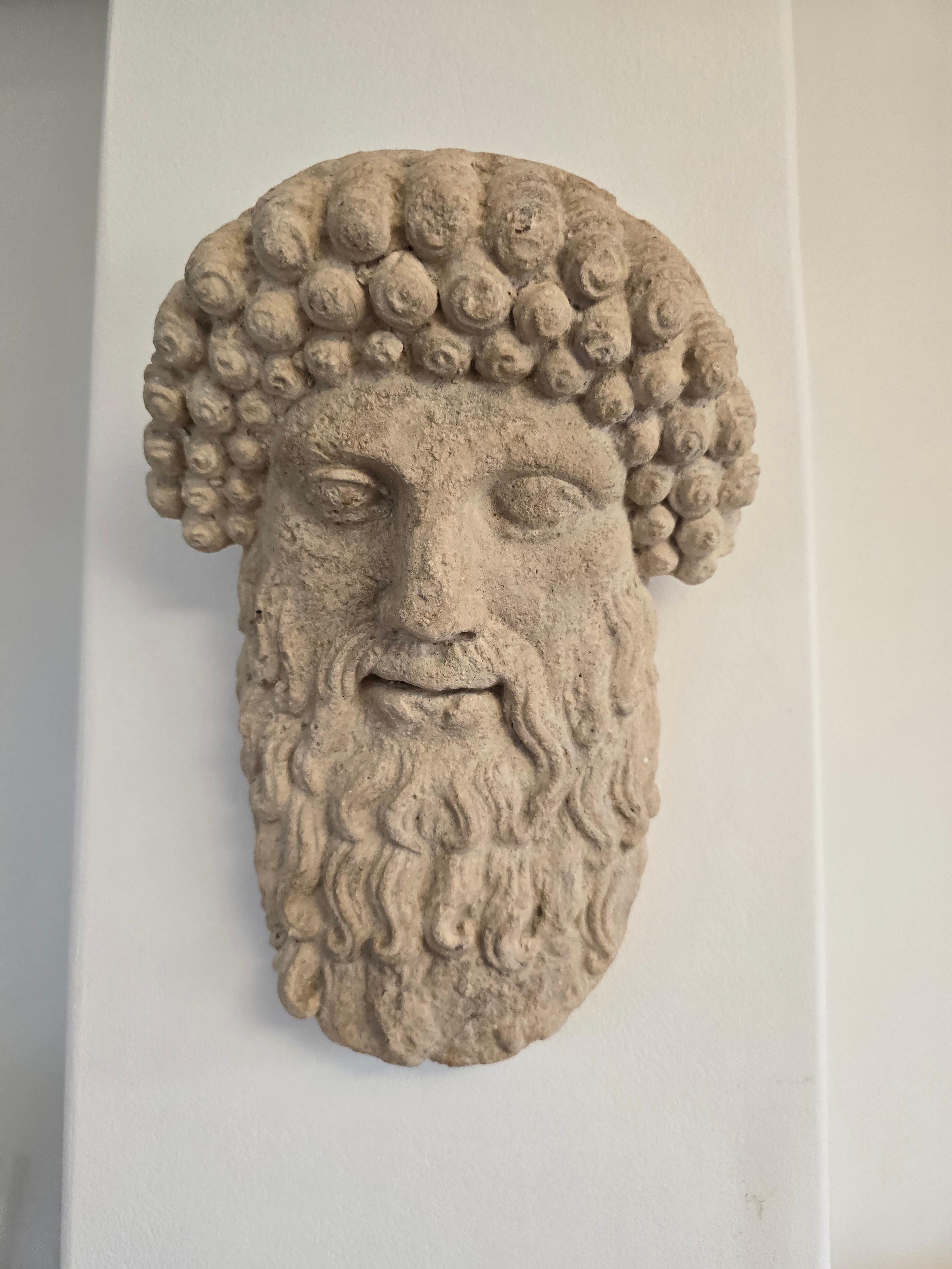 Este notable rostro antiguo de un dios griego, elaborado en terracota o un material similar, es una asombrosa representación del arte clásico. Con sus intrincados detalles y expresivos rasgos, esta pieza no es sólo un elemento decorativo, sino