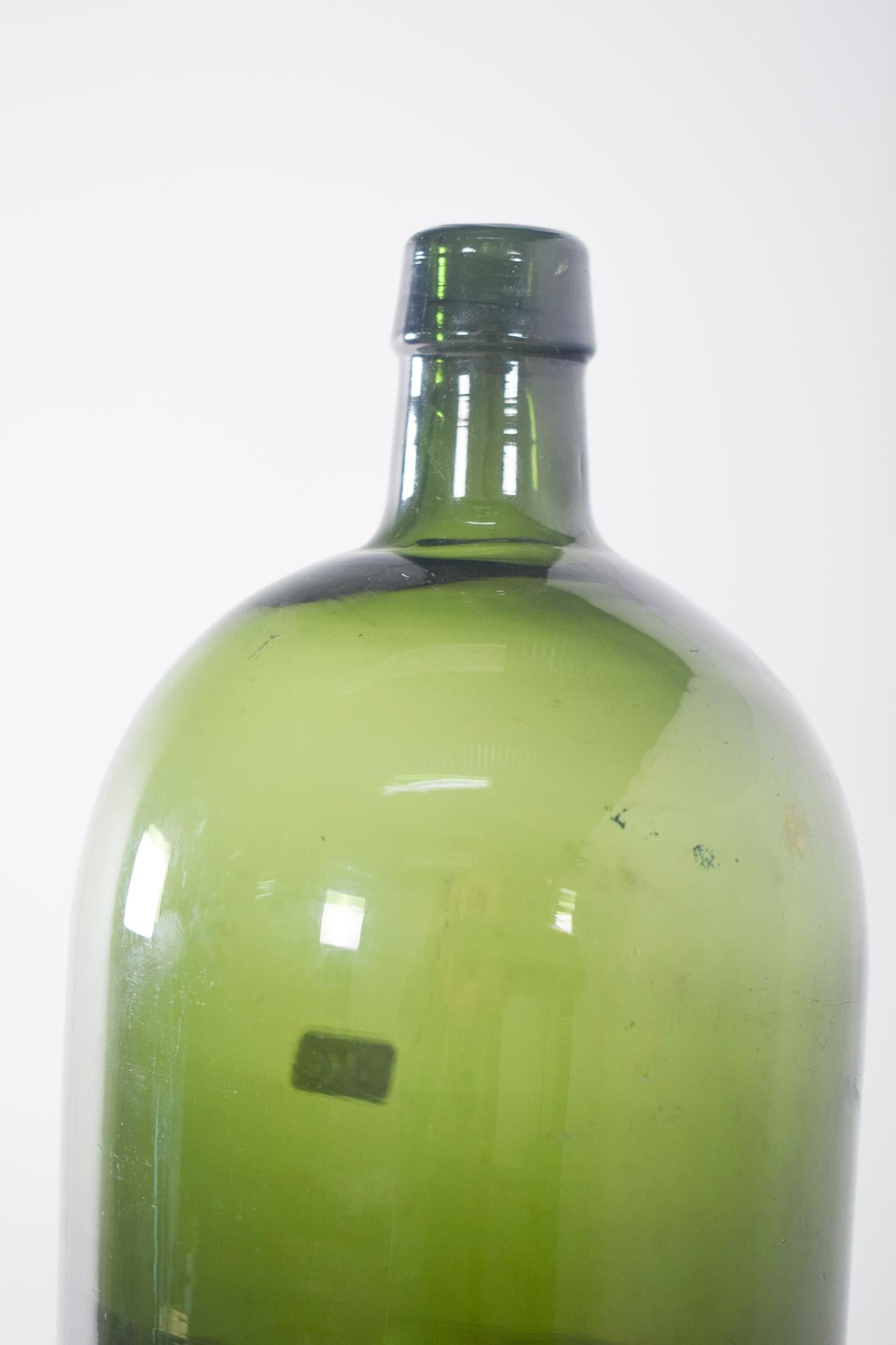 Green glass bottle. Traditionally used to store wine, olive oil or other liquids in large quantities. The bottle is round, with a narrow neck and a smooth finish, characteristic of industrially blown or moulded bottles. The glass has a translucent