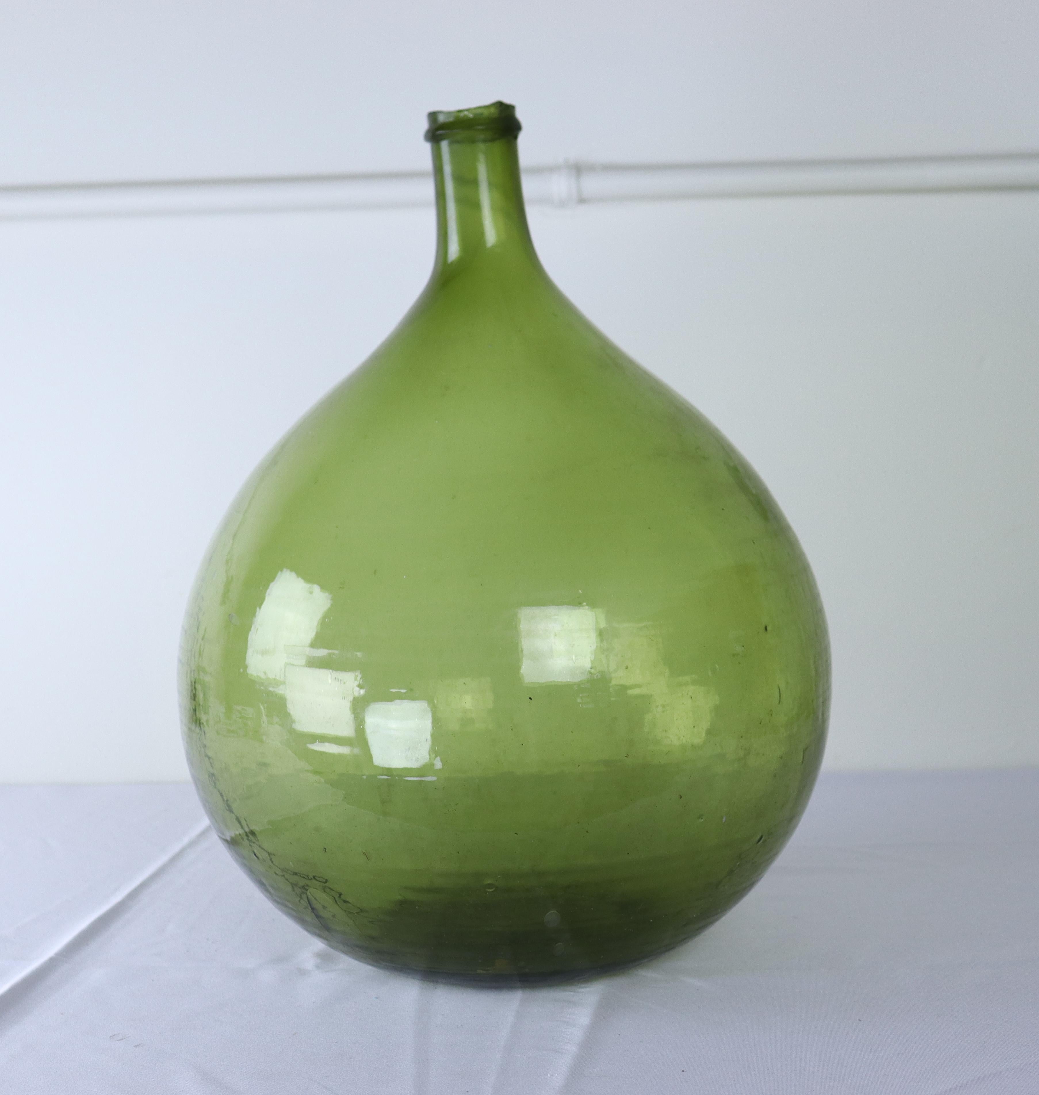 A large olive colored glass demijohn bottle.  These were used from the late 17th Century through the 19th Century to carry wine and spirits. Handblown, each has their own unique characteristics, from the tilt of the gooseneck to the trapped air