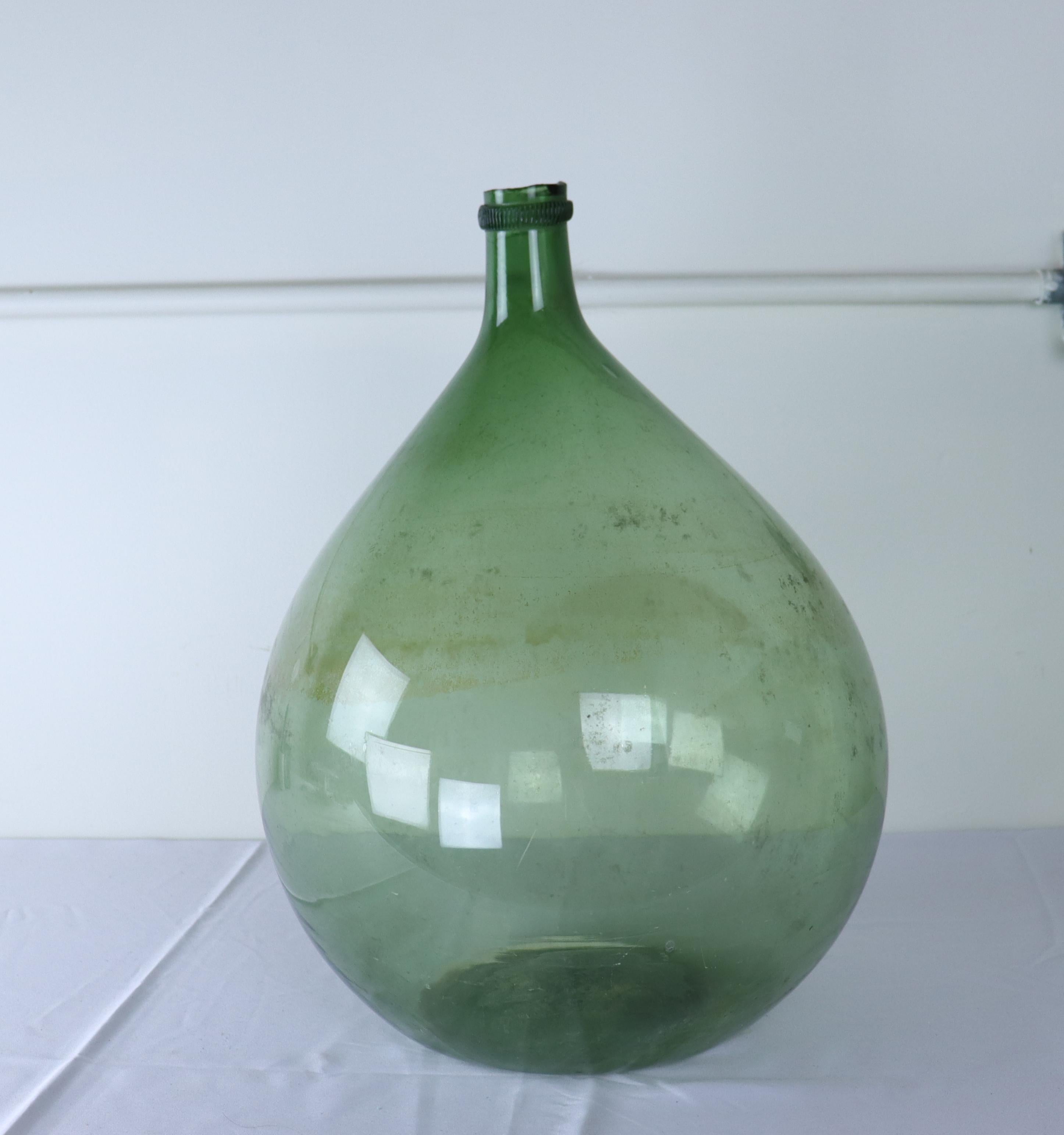 A large olive colored glass demijohn bottle.  These were used from the late 17th Century through the 19th Century to carry wine and spirits. Handblown, each has their own unique characteristics, from the tilt of the gooseneck to the trapped air