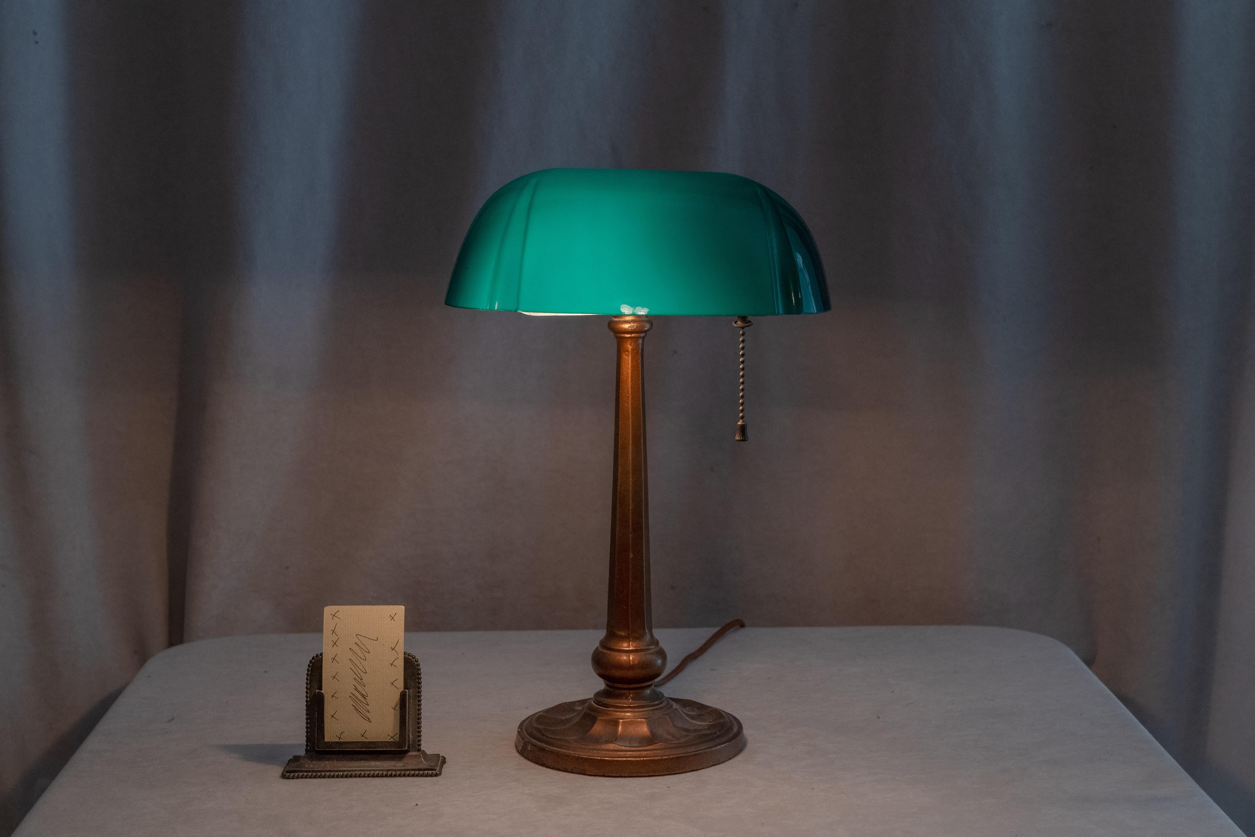  This neat banker's lamp is not your standard fare. Note the work on the shade with the indentations on the front of the shade in 2 places. The base is also not you standard as it is cast and has a pleasing art deco style. I believe lamp is by the