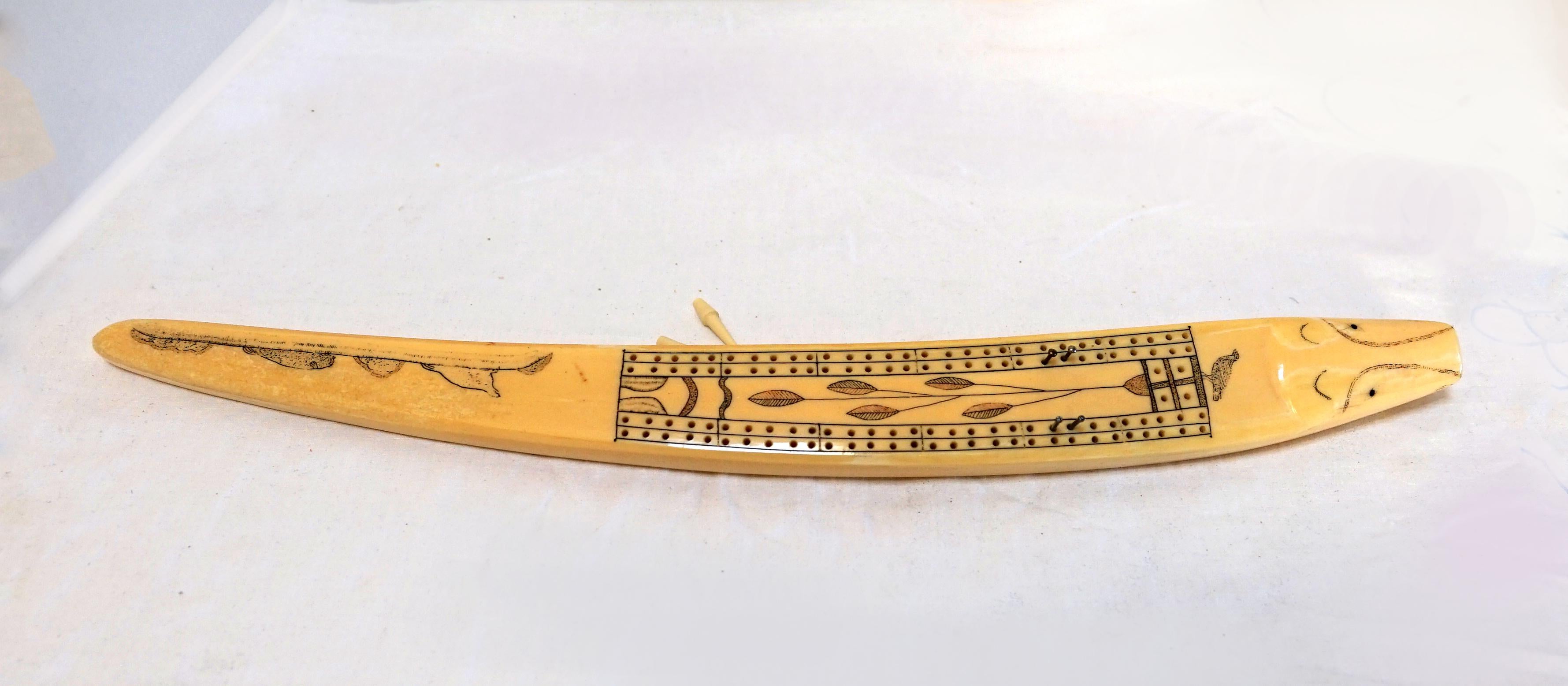 This is a rare and collectible group of three Alaskan scrimshaw articles with a 19th century cribbage board carved from a walrus tusk and two utensils.. 
The Cribbage board is late 19th/early 20th Century.
Comprising an Inuit cribbage board and two