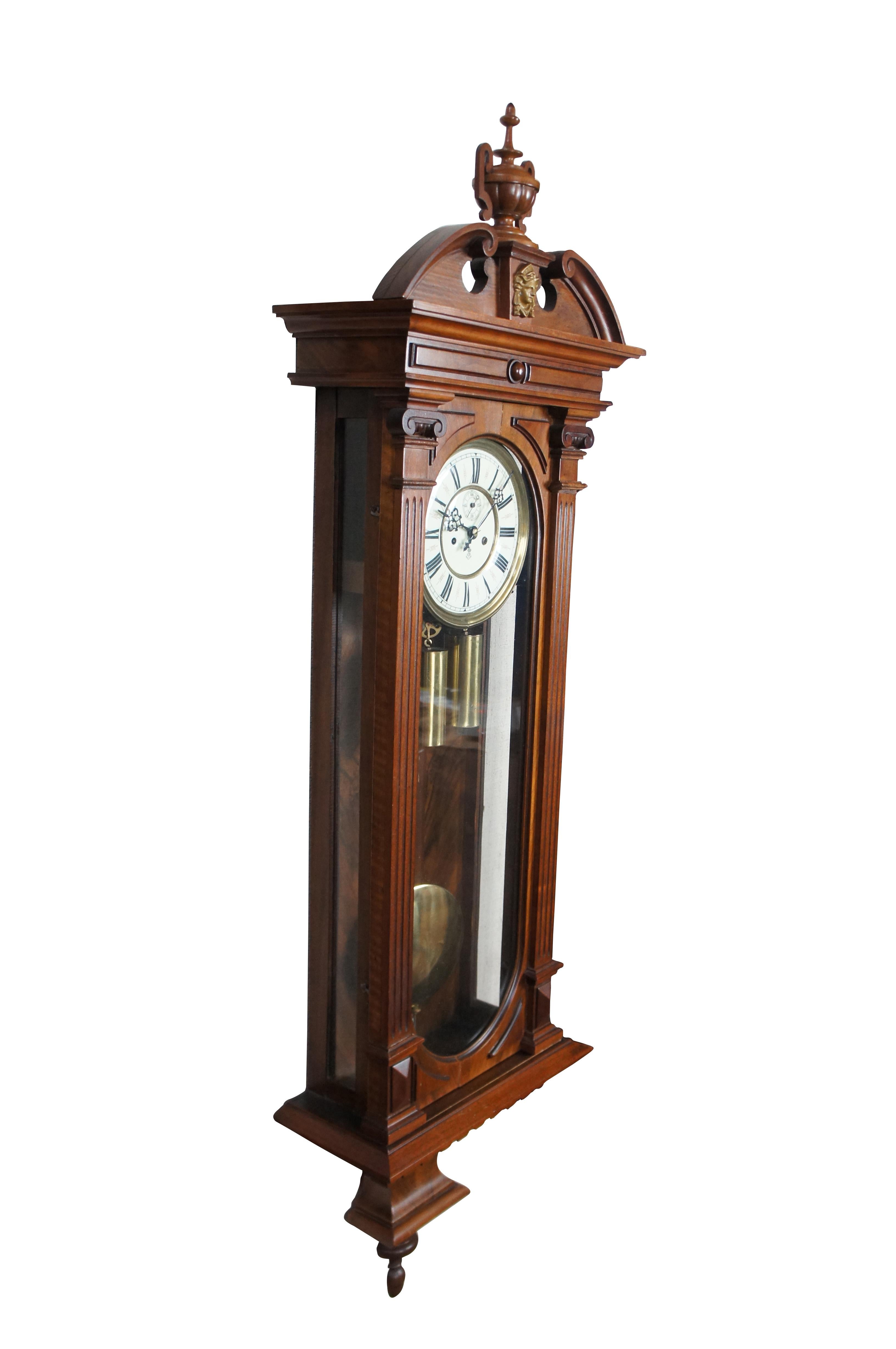 A beautiful antique turn of the century Gustav Becker weight driven Regulator wall clock. Features a long walnut case accented by a central trophy urn over a caryatid mount and a large glass front flanked by Grecian style fluted columns with