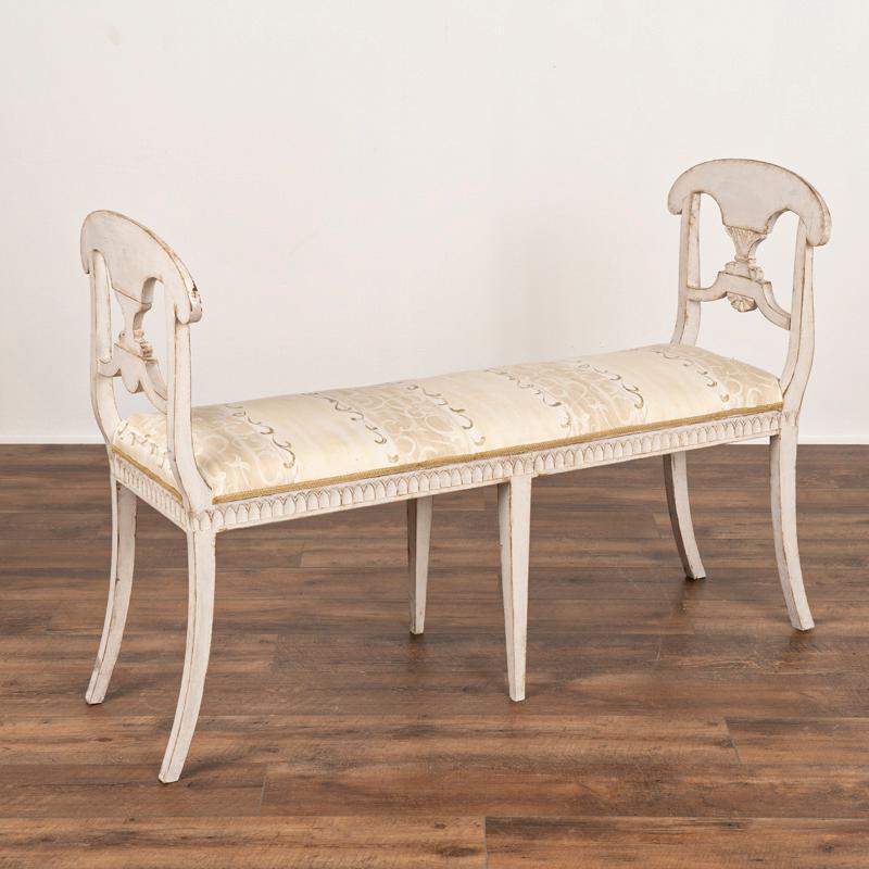 This lovely Gustavian painted window seat or bench displays classic decorative details in the curved arms and carving under the seat. This narrow bench has a newer, professionally applied layered painted finish of antique white and dove gray fitting