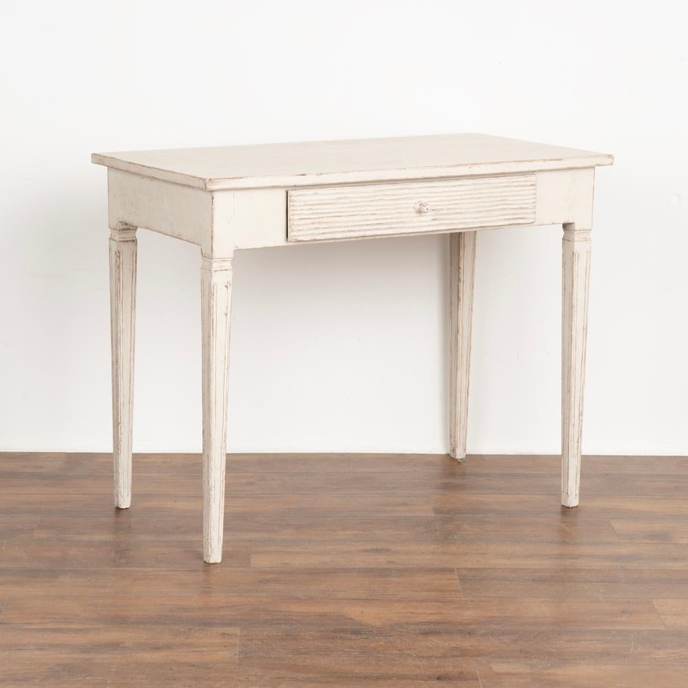 Swedish Gustavian style side table, may also be used as a small desk. 
Traditional tapered legs, horizontal fluted carving along single drawer.
Restored, later professionally painted in layered shades of white. 
Any old nicks, scratches, dings,