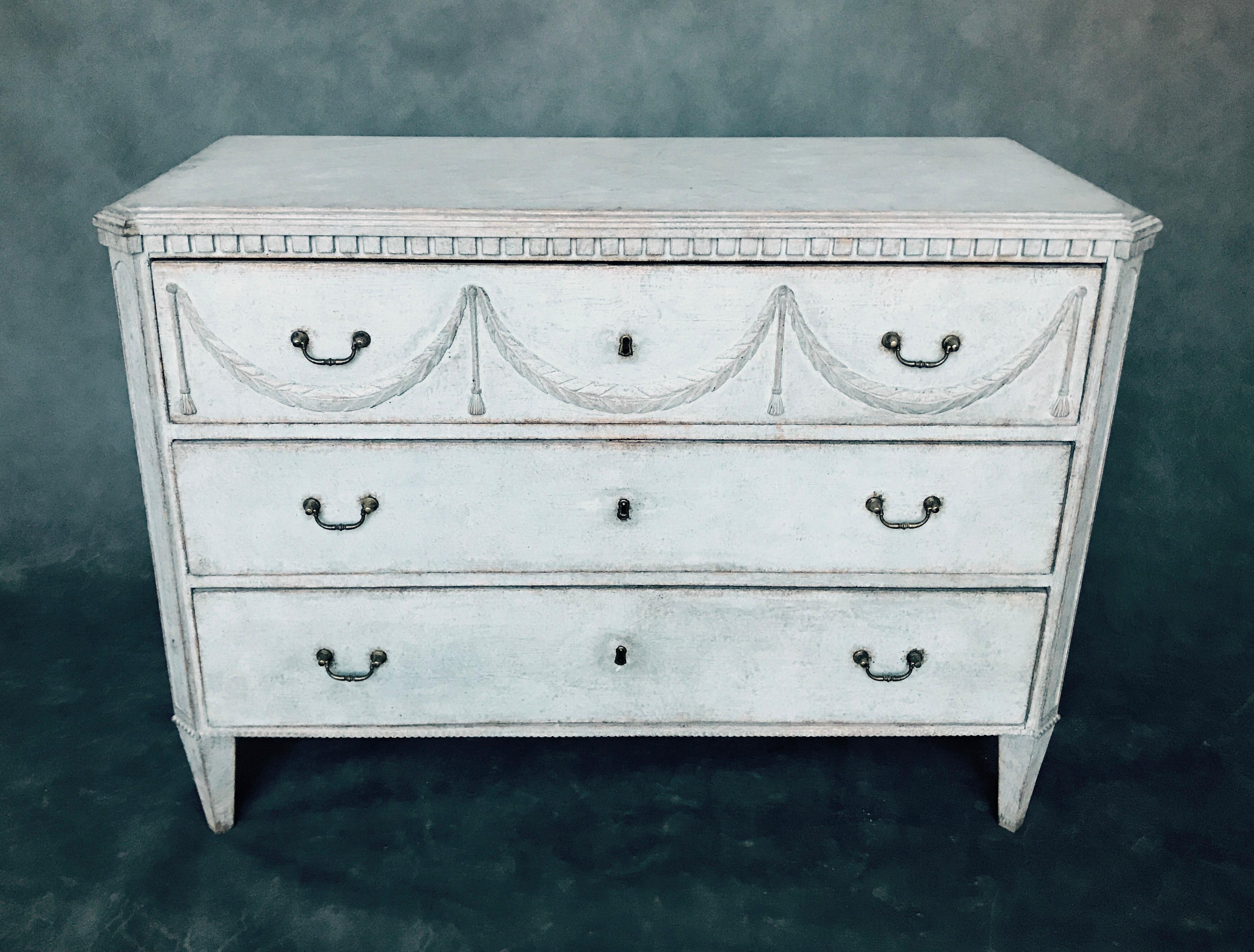 Antique chest with three drawers in the Gustavian style. In the upper part of the furniture there are beautiful decorations, wreaths. This furniture is in light gray color and the top is painted marble.