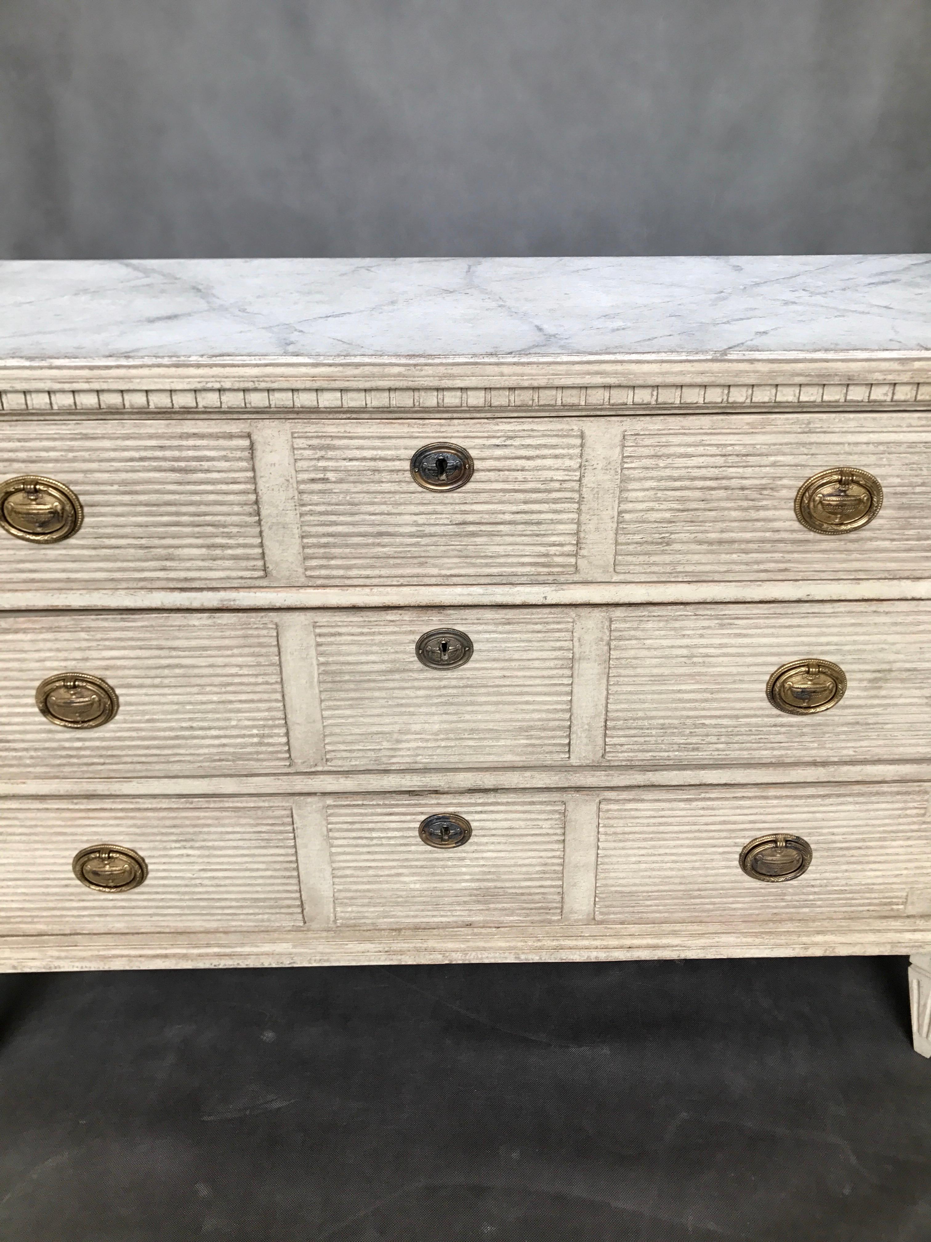 Beautiful, wide in the Gustavian style chest with three-drawers in cream color. On the front there are unique horizontal indents. The legs are tapered down and have vertical cutters. The top is painted marble. Age, circa 1840.
  