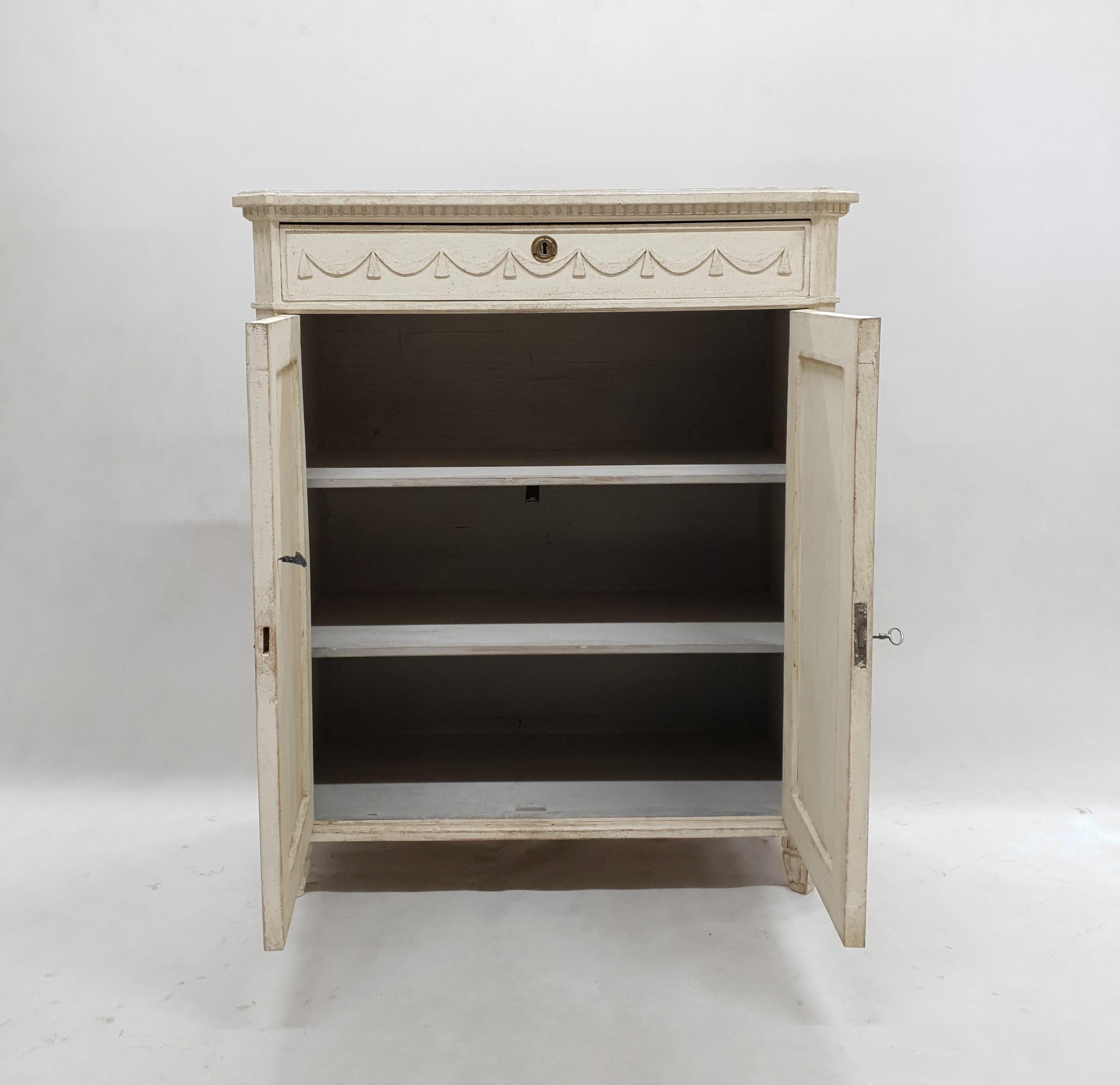 This Gustavian sideboard features one drawer and two doors. On the drawers there are delicate carving in the shape of wreaths. On the doors are vertical cutters. The exterior is in an antique white color and the interior is light grey.
 
