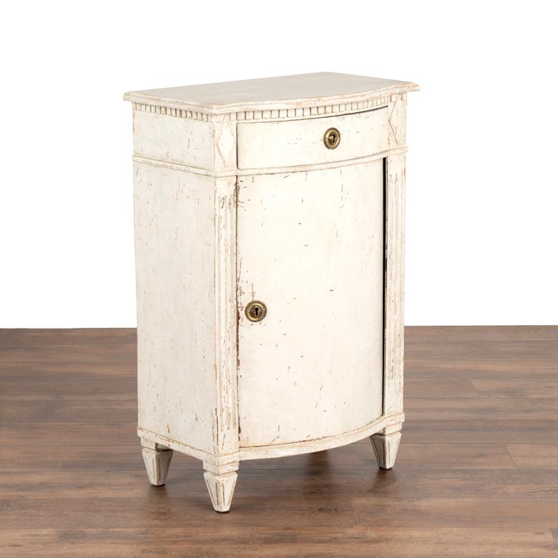 Gustavian style small cabinet/nightstand.

This small case piece features a molded edge top with carved dental molding frieze above curved cabinet door.
Raised on four tapered fluted feet.
Restored, antique conservator painted in white with