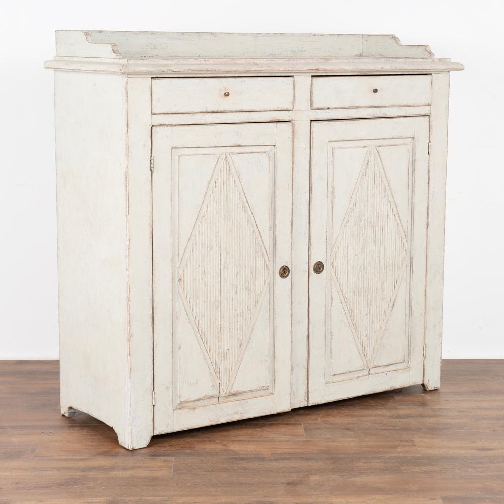 Gustavian white painted tall pine sideboard or server cabinet.
Traditional decorative fluted diamond panels on cabinet doors, two small upper drawers with small brass pull.
One key included to use as pull for doors. 
Interior holds three shelves