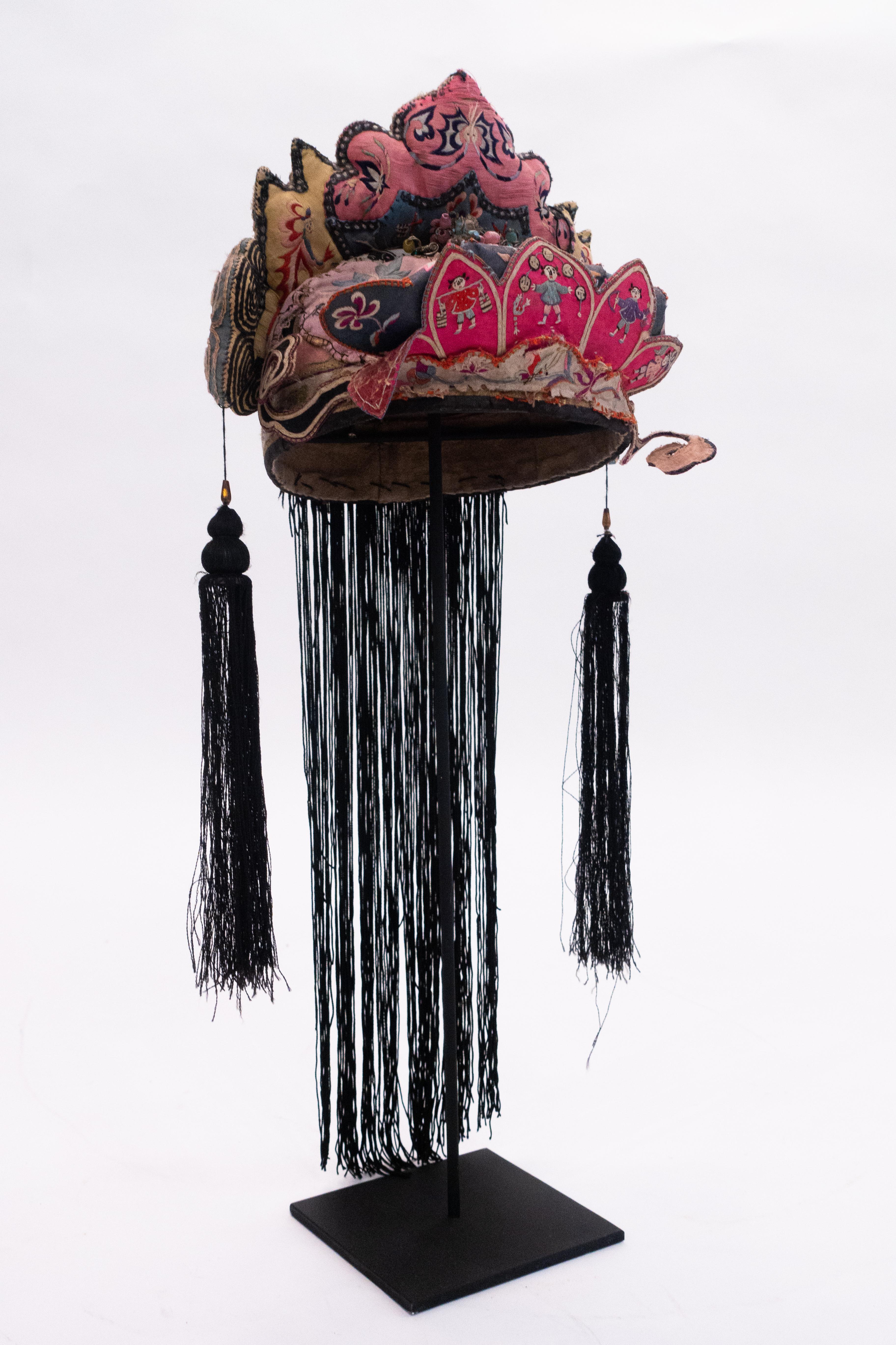 Handbestickter Kinderkopfschmuck aus Seide vom Stamm der Miao-Minorität mit Perlen und Quasten aus dem frühen 20. Jahrhundert auf einem speziell angefertigten schwarz lackierten Metallgestell.

 