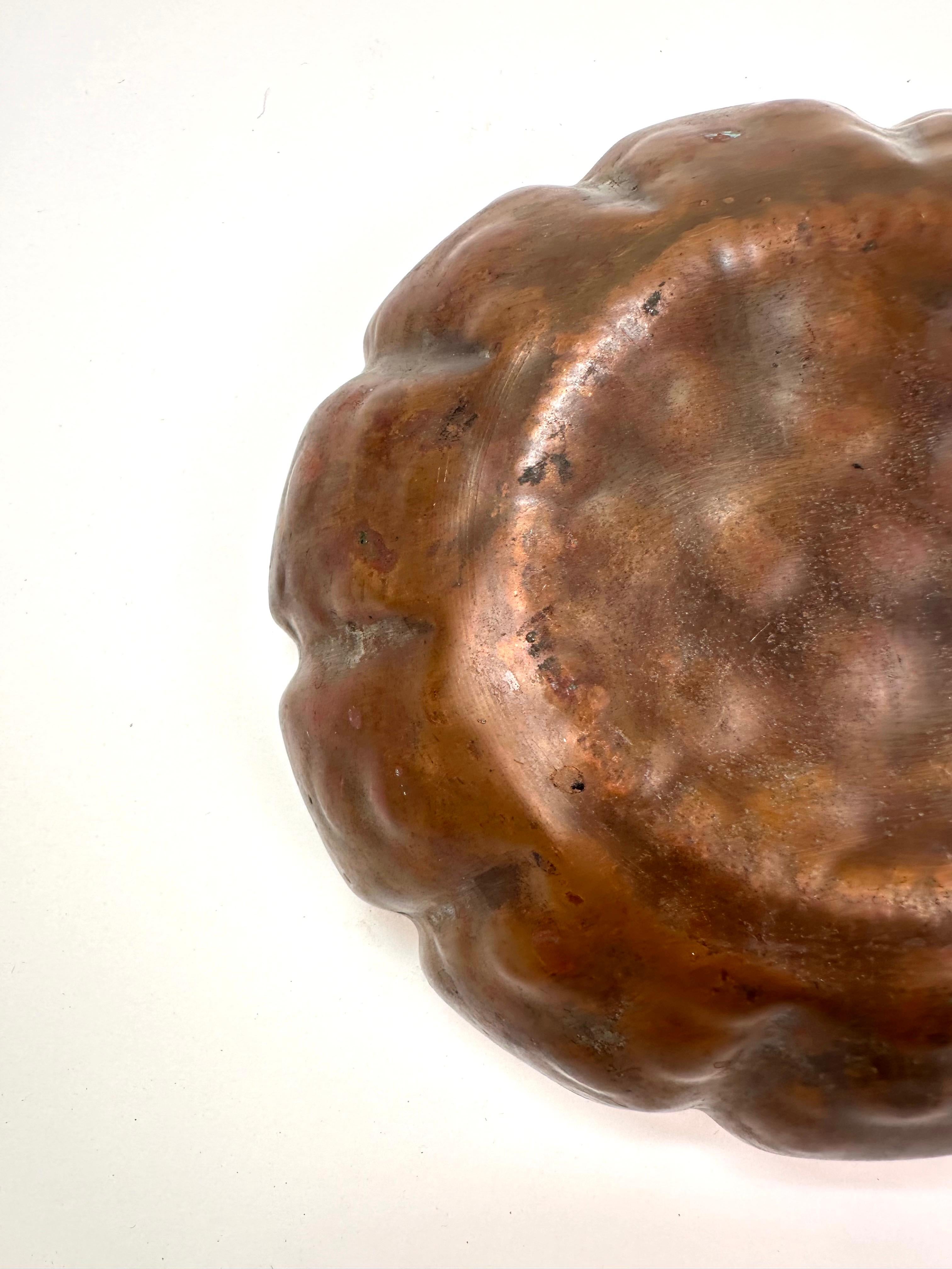 Una ciotola in rame antico splendidamente invecchiato con un design martellato a mano e bordi smerlati e scanalati. La patina naturale e le macchie di verderame sulla superficie ne esaltano il fascino vintage, evidenziandone l'età e l'autenticità.