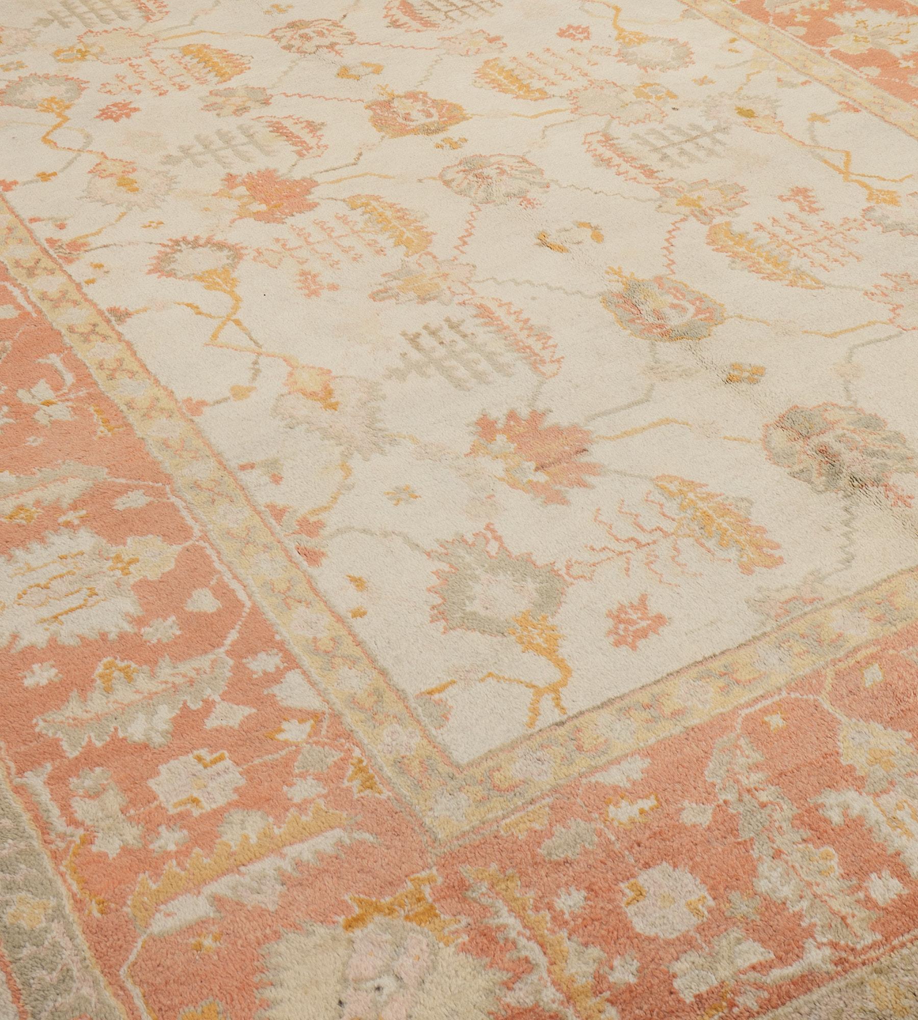 This antique Oushak rug features an ivory field with an overall design of soft terracotta-pink, sand-yellow and soft-grey palmette and serrated leaf vine, in a broad soft terracotta-pink border with large ivory palmettes linked by angular floral