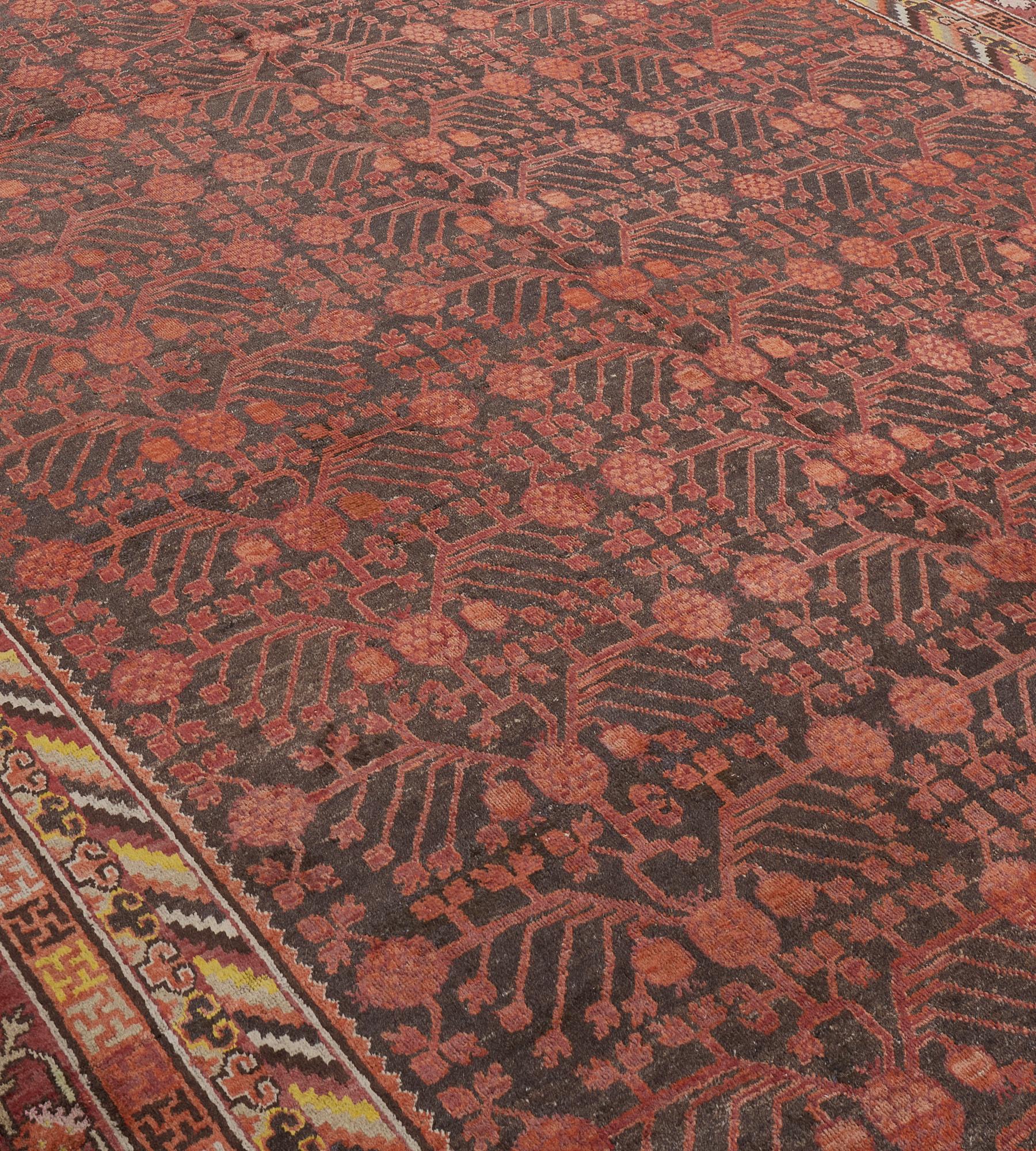 This antique, circa 1900, Khotan rug has a mole-brown field with an overall dense terracotta-red pomegranate vine forming three columns issuing angular floral vine stems, in a narrow border of linked T-shaped motifs forming polychrome open squares