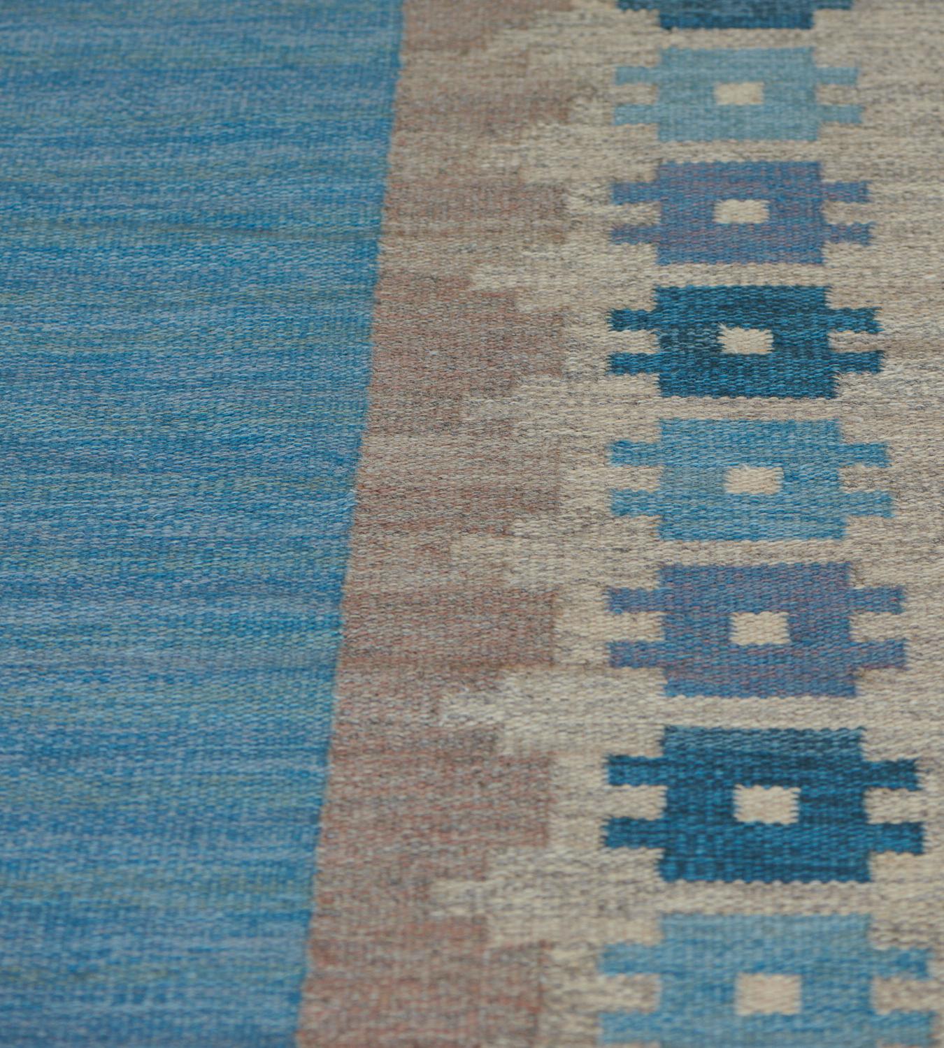 This antique Swedish rug features a plain shaded sky-blue field, in a broad border of shaded sandy-brown outward facing part stepped lozenges with a row of shaded blue bold square motifs issuing a pair of minor panels at each side, signature initial