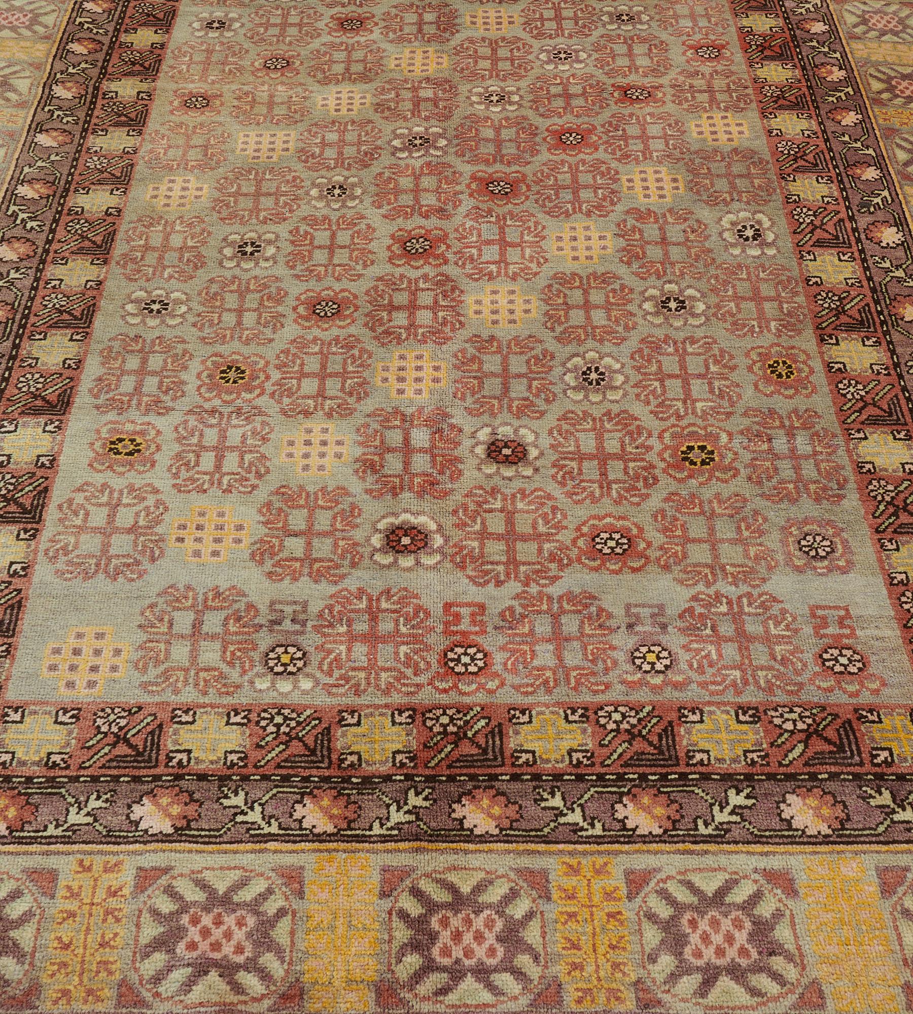 This antique Khotan rug has a pale blue field with diagonal rows of hooked terracotta-red floral motifs alternating with flowerheads and sandy-yellow stepped lozenges, in a pale blue border with hooked square panels divided by sandy-yellow floral
