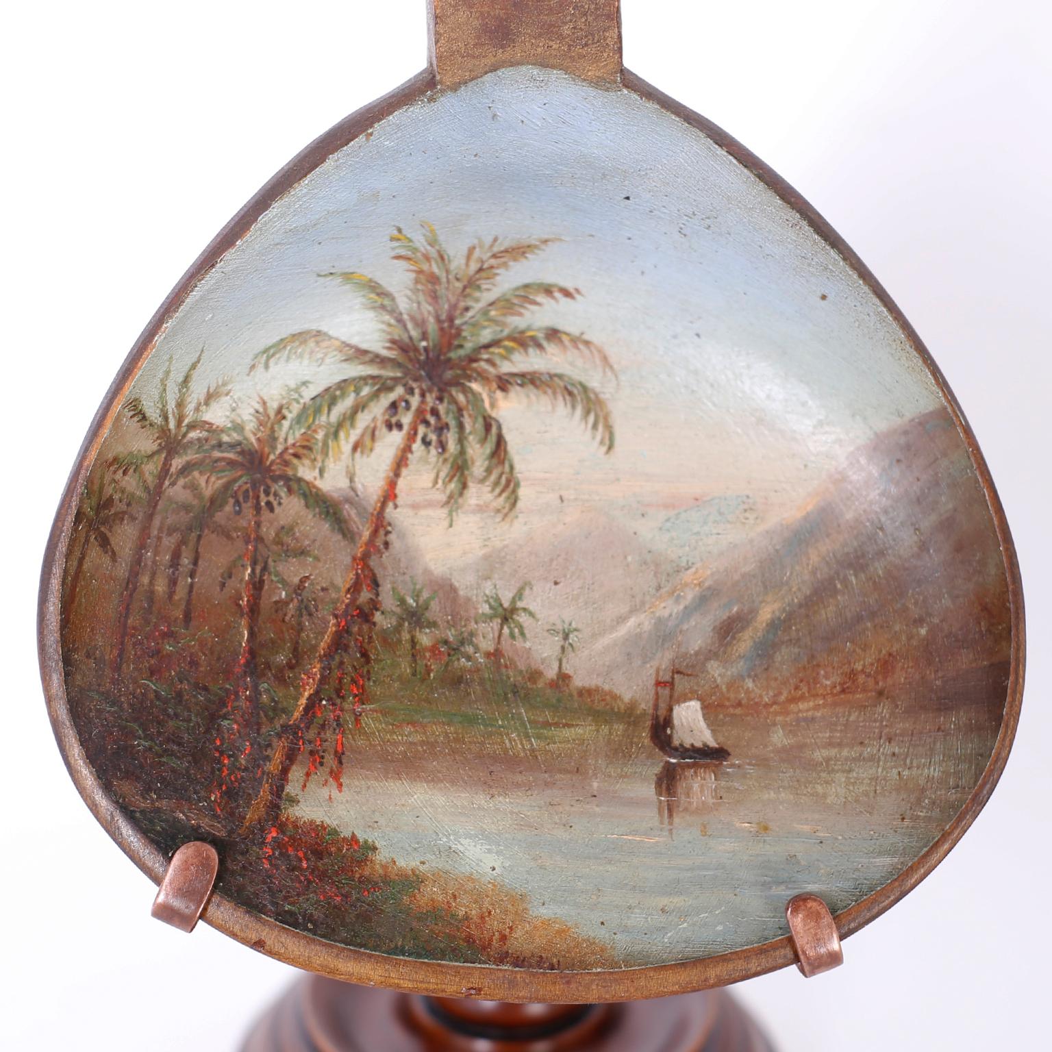 Rare and unusual antique grain scoop crafted in indigenous hard wood and expertly decorated with a sophisticated tropical oil painting of palm trees, mountains, water and a sail boat, mounted on a custom stand.