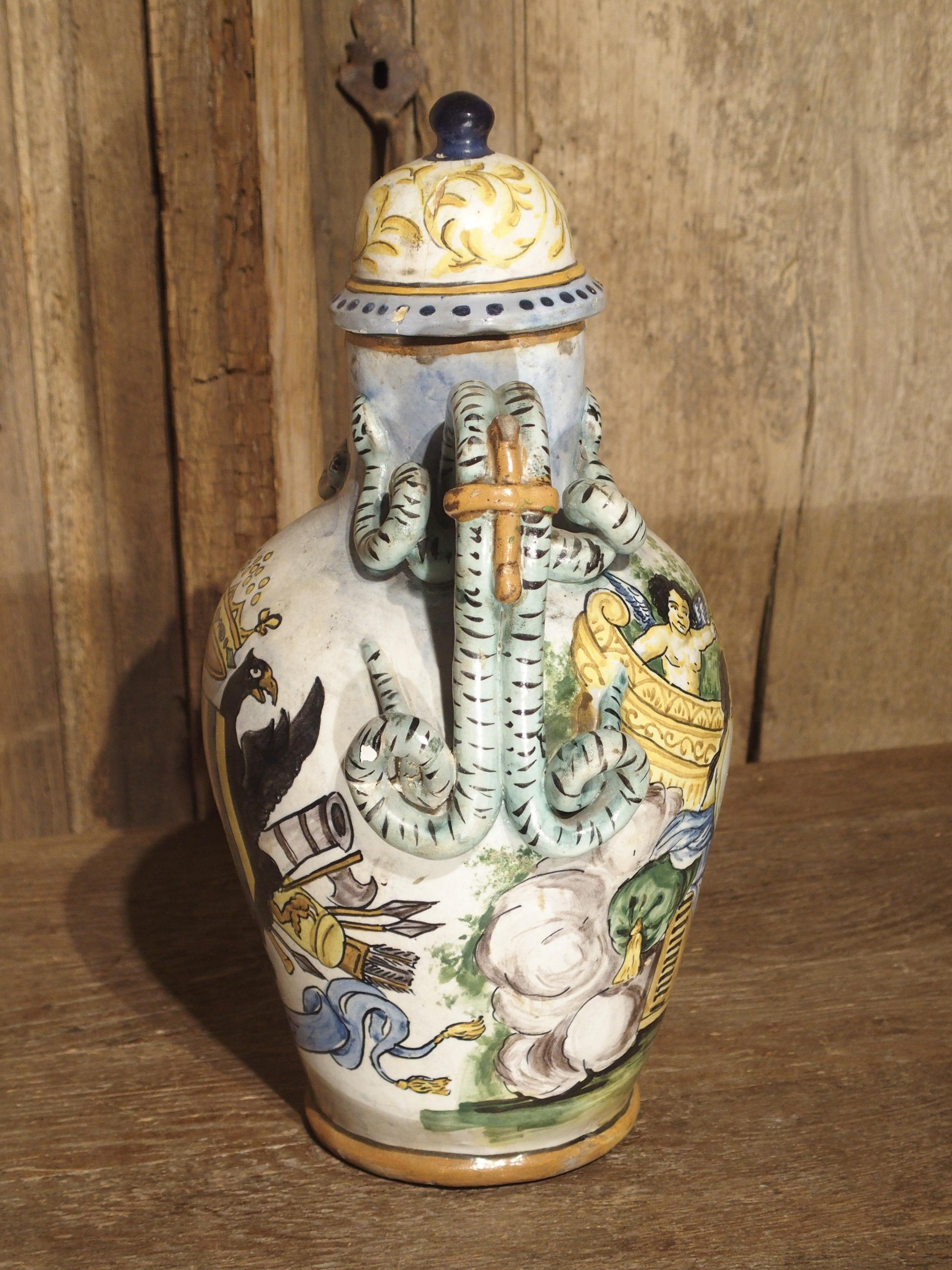 From Italy, this hand painted majolica vase has two scenes divided by scrolling snake handles. One side depicts a trophy of arms with black eagle supporters, a flag and a ribbon dated 1745. The other side depicts a figural scene with playful dogs on